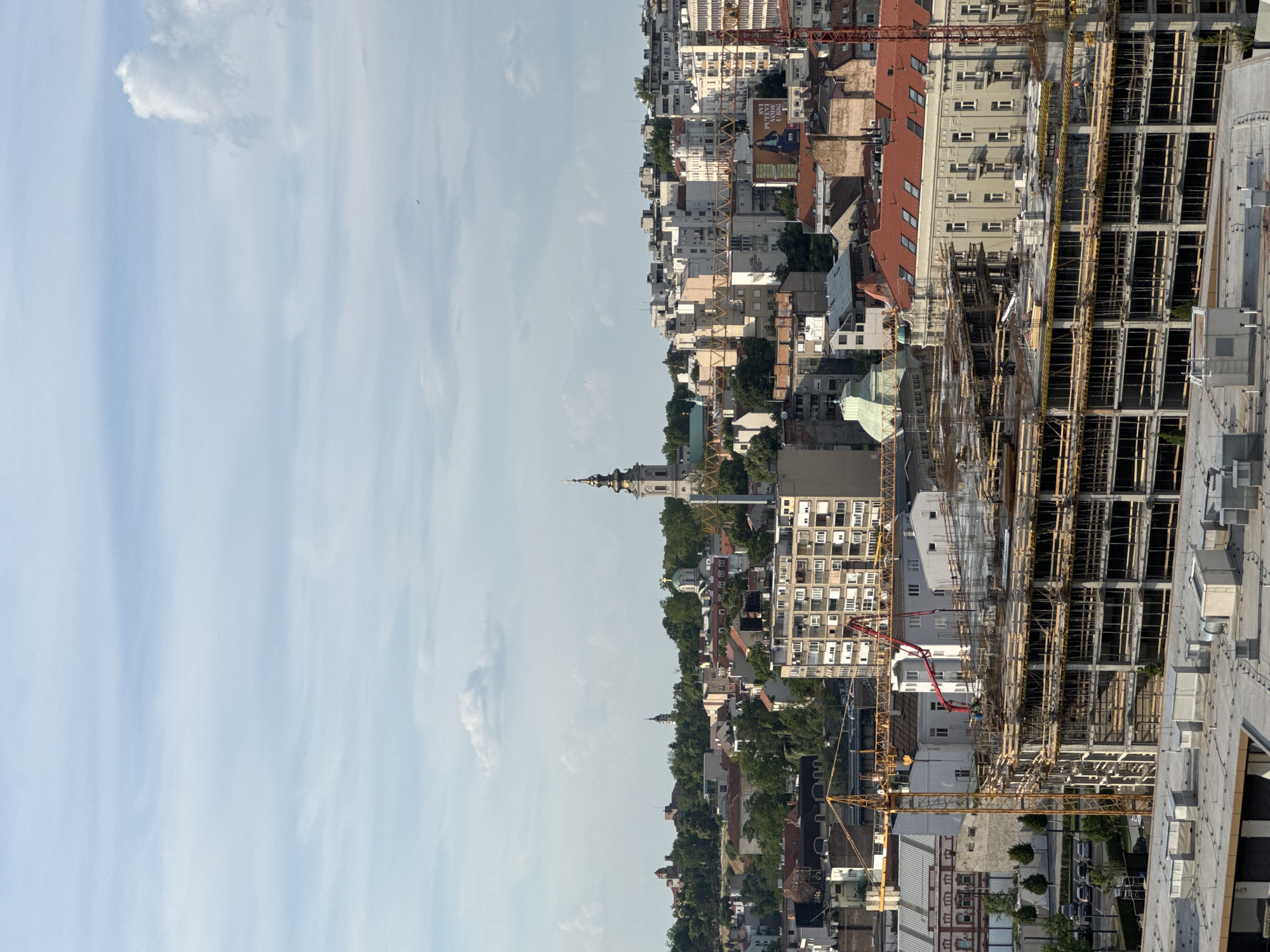 The image shows a cityscape in Belgrade, Serbia. The scene is oriented vertically, with the sky on the left and the urban landscape on the right. The sky is a light blue with wispy clouds. The urban landscape features a mix of old and new buildings, including residential structures, commercial properties, and a prominent church spire. Several construction cranes are visible, suggesting ongoing development. The buildings range in height, with older structures displaying pitched roofs and newer high-rise buildings under construction. The lighting is bright, indicating a sunny day. There are no people visible in the image.