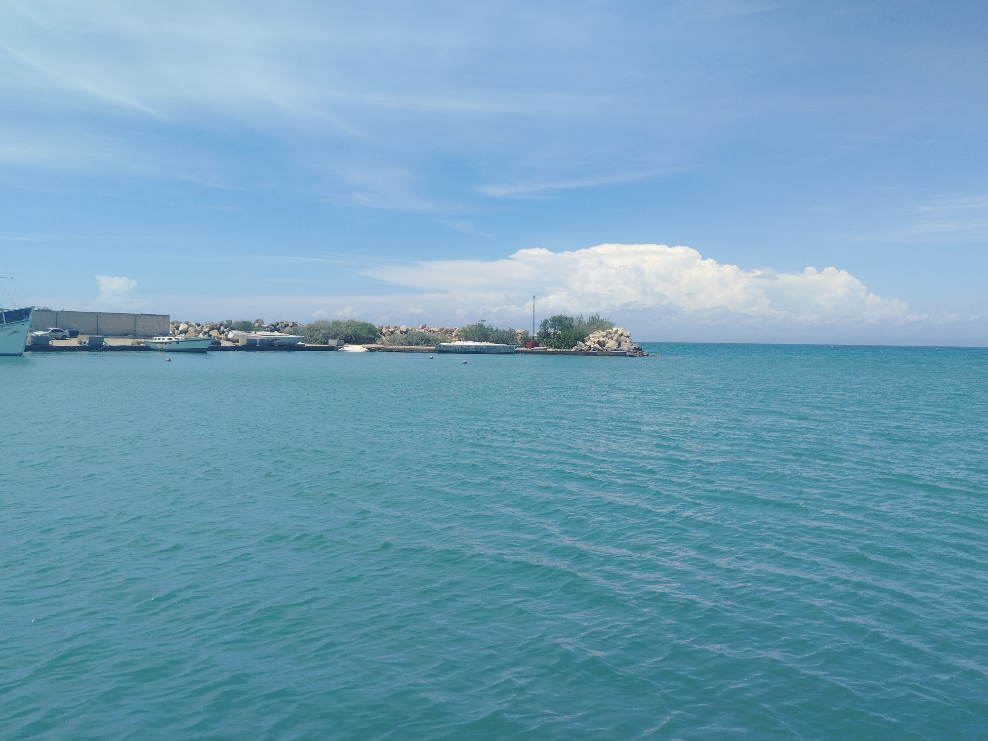 This media file shows a view of a calm, turquoise sea with a clear blue sky filled with wispy white clouds. In the distance, along the shoreline, there are several boats docked and moored. Some of these appear to be sailboats and smaller vessels. The shoreline itself is lined with a concrete structure and a rocky embankment, with some vegetation visible.
The scene suggests a peaceful day at a harbor or marina. The lighting and the bright sky indicate that it is likely daytime, possibly late morning or early afternoon. The weather appears to be fair and sunny. The presence of the docked boats implies a location related to maritime activities, such as a port or a coastal town. The overall mood of the image is serene and tranquil, reflecting the calm water and the clear sky. There is no visible text in the image.