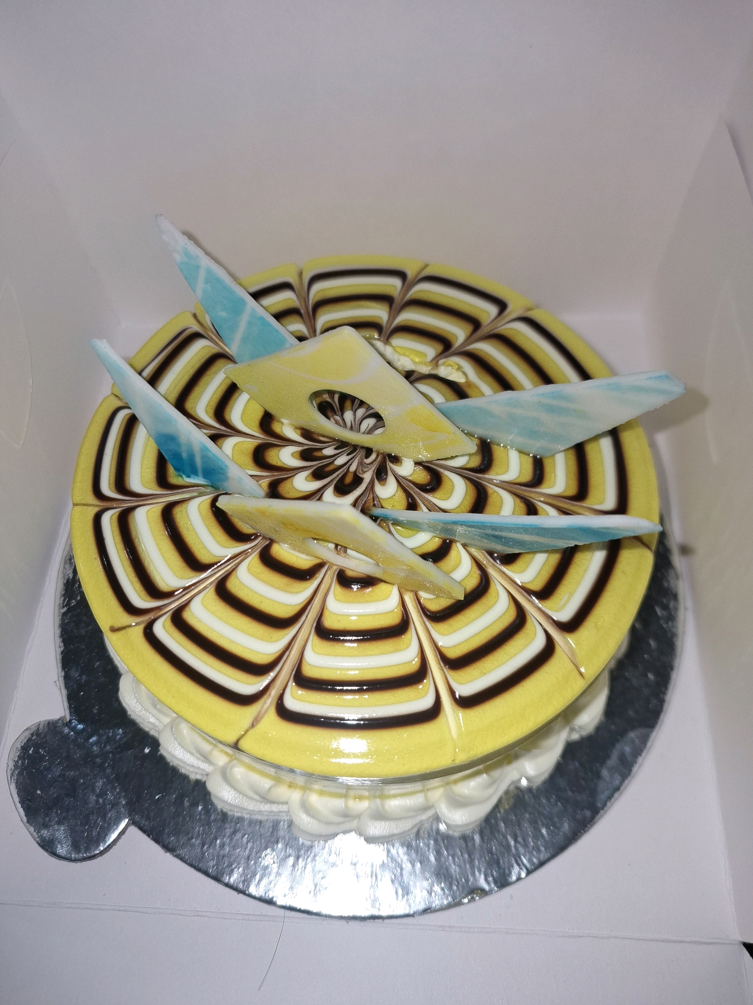 A round, yellow cake with a swirled chocolate and white icing design is presented in a white box. The cake is garnished with three light blue, pointed decorative pieces and two yellow, leaf-shaped decorations. The cake sits on a silver cake board, which is partially visible within the box. The image is taken from a high-angle perspective. The exact location of the cake within the city of Yamunanagar, India is not discernible from the image.
