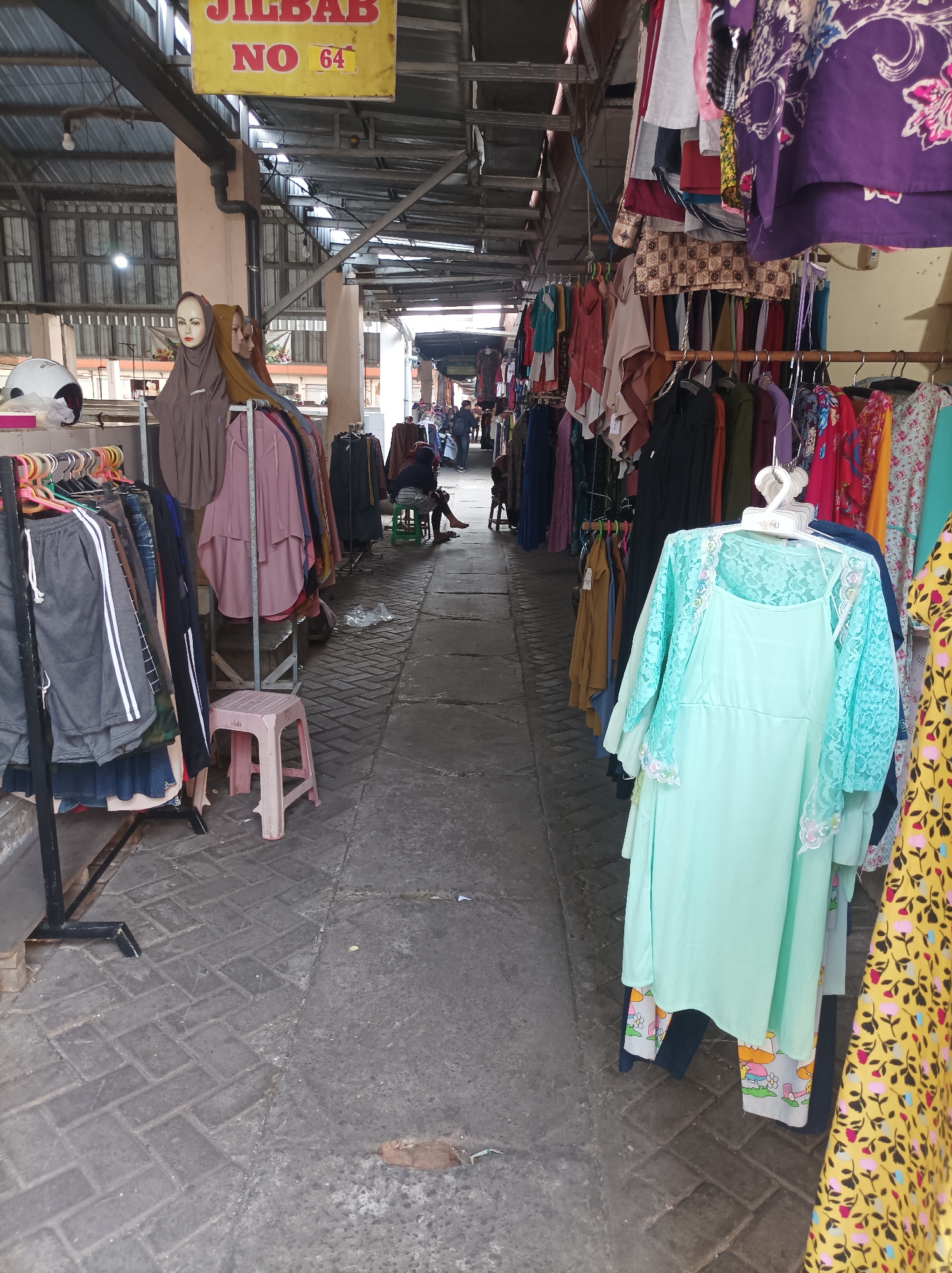 The image depicts a market stall in Nganjuk, Indonesia, likely located in a covered outdoor or semi-outdoor area, possibly part of a larger traditional market or bazaar. The stall features various clothing items for sale, including a mannequin wearing a brown hijab, several other hijabs and modest clothing on display, and racks of shirts and dresses. In the foreground, a light green dress with lace detailing is prominently displayed. To the right, a yellow dress with a floral pattern is visible. The ground is paved with interlocking bricks. In the background, a few people are seen browsing or walking through the market. A yellow sign with red lettering reading "JILBAB NO 64" is hung above the stall. The overall scene suggests a bustling commercial environment focused on clothing retail.