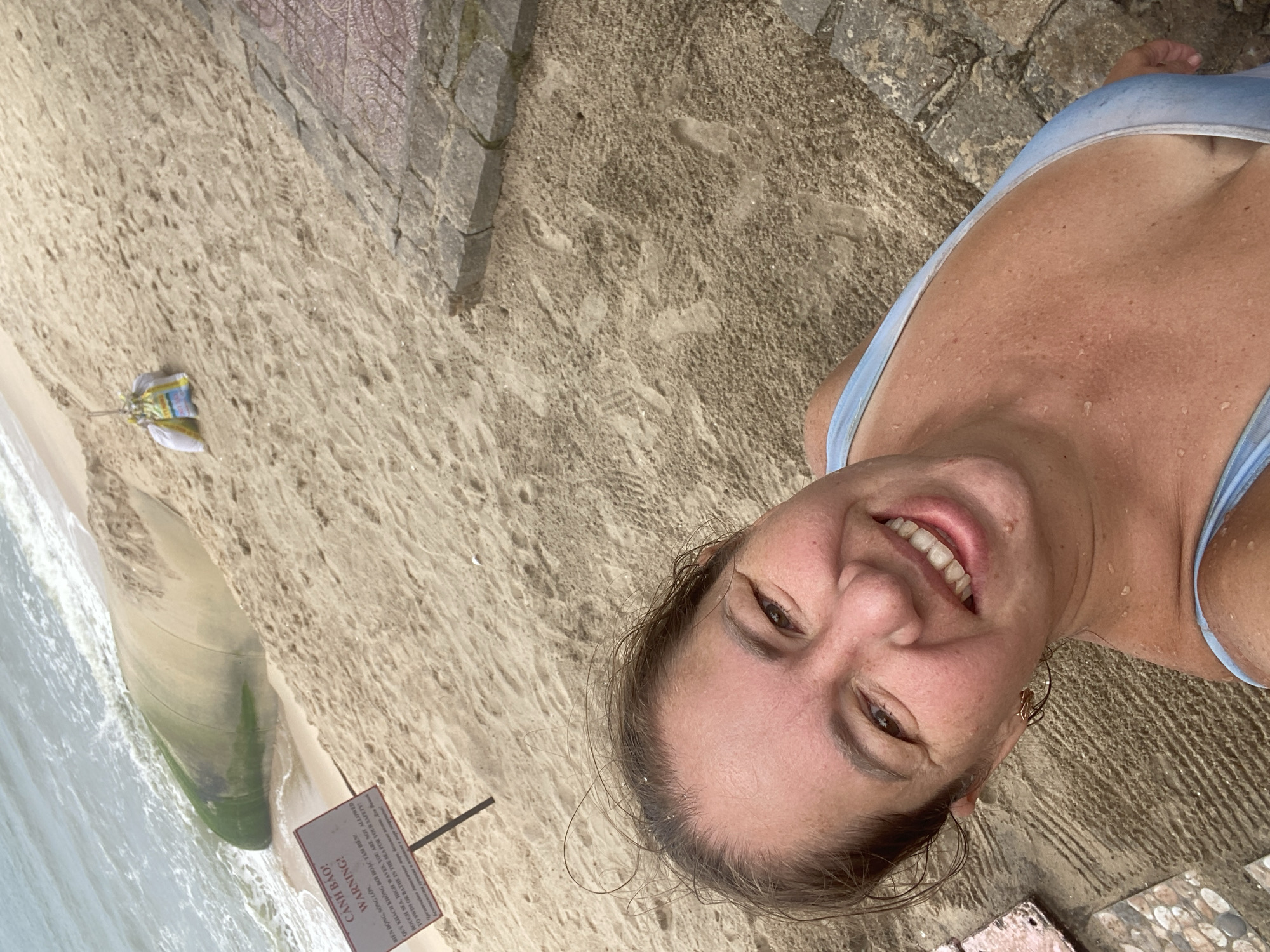 A woman with wet hair and skin is taking a selfie on a sandy beach in Mũi Né, Vietnam. She is wearing a light blue swimsuit and smiling at the camera. The background shows a sandy incline leading down to the ocean, where waves are crashing. There is a large, curved object partially buried in the sand near the water, possibly a discarded piece of equipment or debris. A red and white warning sign is planted in the sand, with text in multiple languages, indicating rough seas and a warning against swimming. Further up the beach, there are two bags filled with sand, likely used for erosion control or construction. To the right of the image, a stone-tiled walkway or steps are visible. The overall scene suggests a potentially hazardous beach environment.
