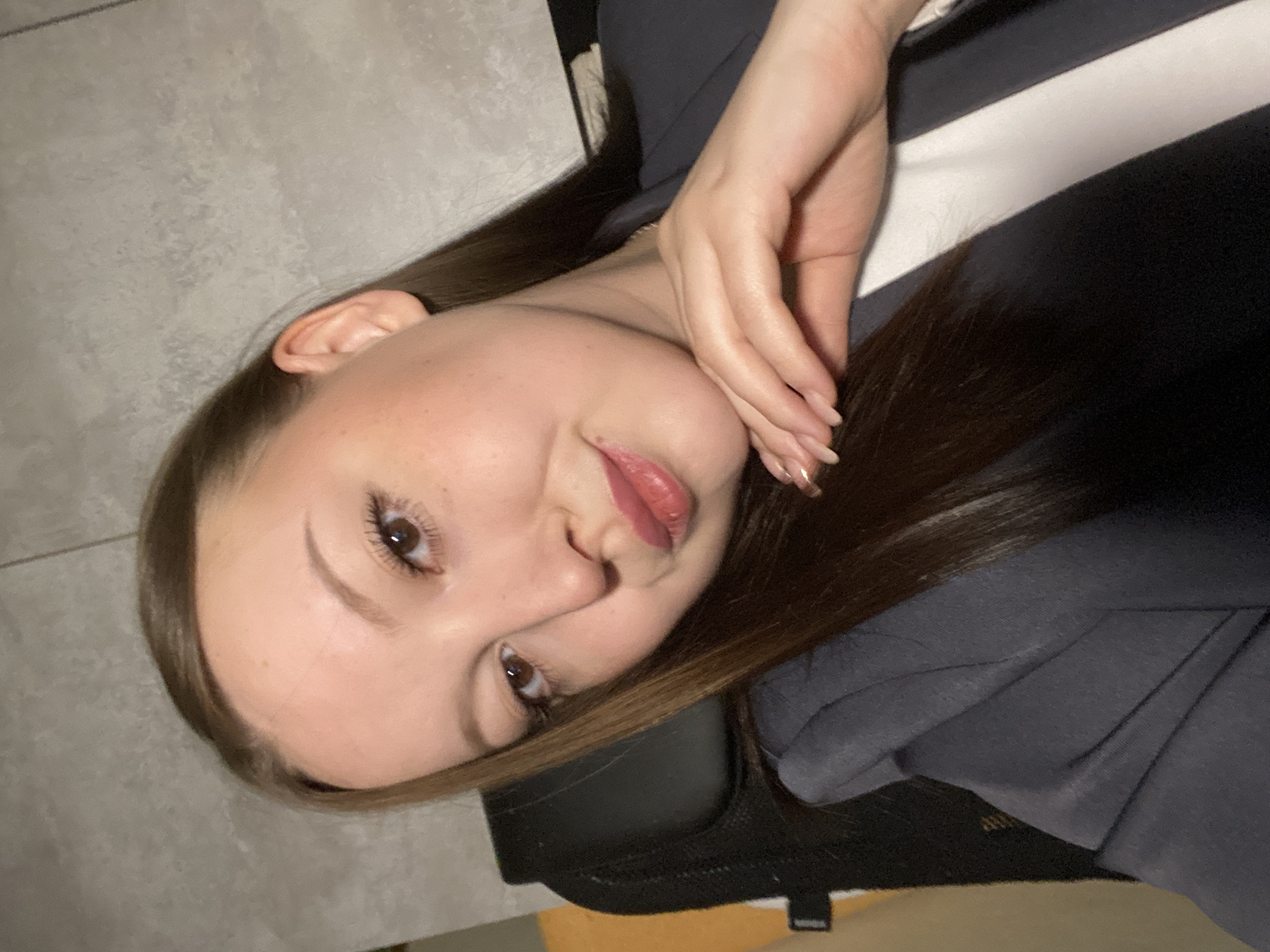 The image presents a close-up, self-portrait style photograph of a young woman taken indoors in Chaikovsky, Russia. Her head is tilted significantly, with her gaze directed towards the viewer, and her right hand is gently placed near her jawline and neck.
The woman has long, dark brown hair that falls over her shoulder. Her complexion is fair, and she has brown eyes with noticeable mascara on her eyelashes and well-defined eyebrows. Her lips are slightly parted, revealing a pinkish-red lip color, and her expression conveys a subtle, almost playful or confident smirk. Her visible hand features long, manicured nails, possibly with a light or French tip polish.
She is dressed in a dark-colored garment, possibly a jacket or hoodie, with a distinctive white stripe visible on the upper right side of the frame. The background consists of a plain, light grey wall with a subtle texture, indicating an indoor setting. A dark object, which appears to be part of a chair or furniture, is partially visible beneath her. The lighting is soft and even, typical of indoor illumination. There is no visible text in the image.