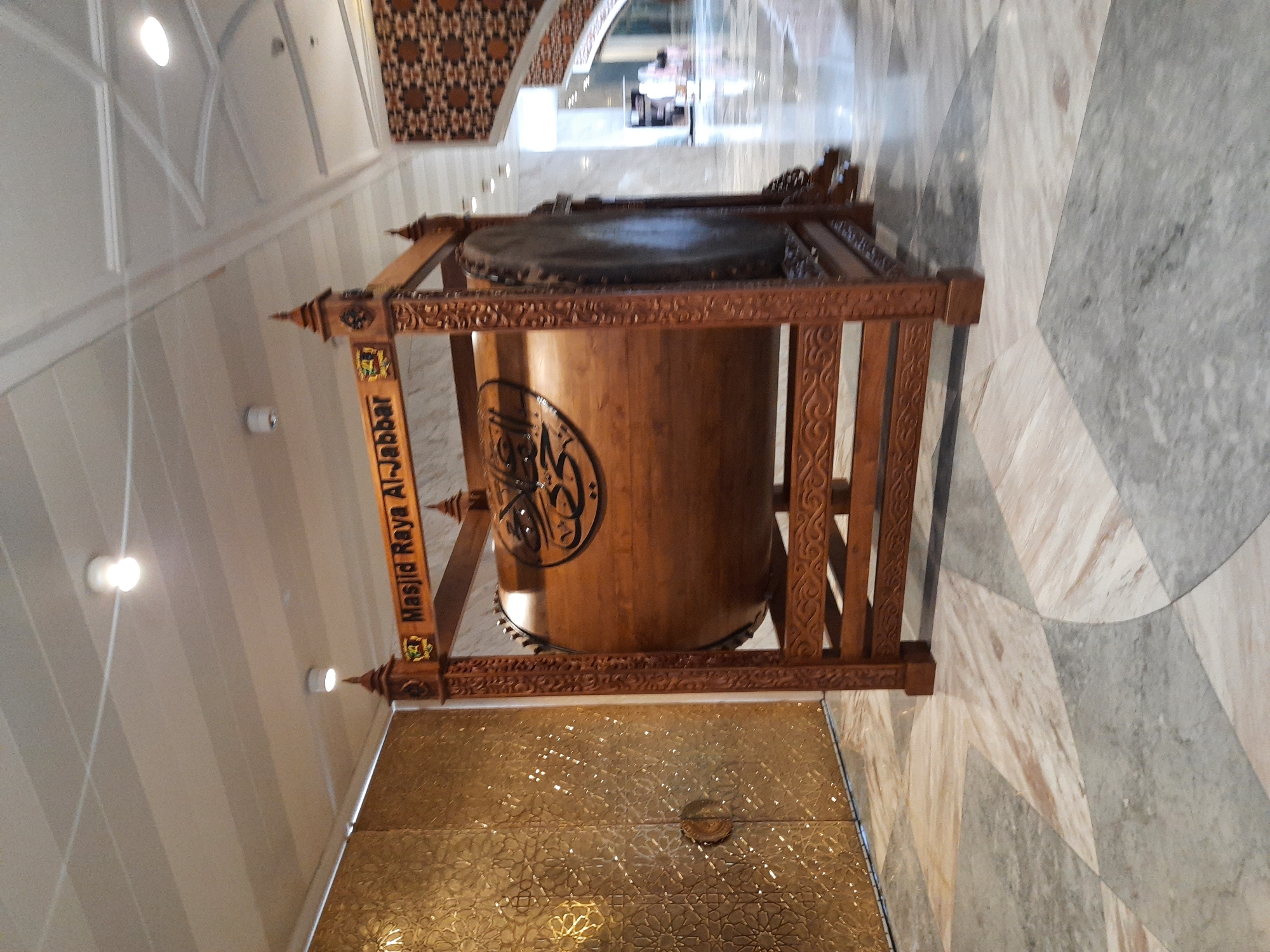 The image shows a large wooden drum, known as a bedug, displayed on a wooden stand in what appears to be the interior of a mosque or a similar public building. The drum has Arabic calligraphy on its surface, and the stand is ornately carved. Behind the drum, to the left, is a large, ornate golden door with a patterned design. To the right of the drum, there is a marbled floor and in the background, a patterned wall and a staircase can be seen. The ceiling has recessed lighting. The text on the drum stand reads "Masjid Raya Al-Jabbar". Based on this text, the location is the Grand Mosque of Al-Jabbar in Bandung City, Indonesia. No people are prominently visible in the foreground.