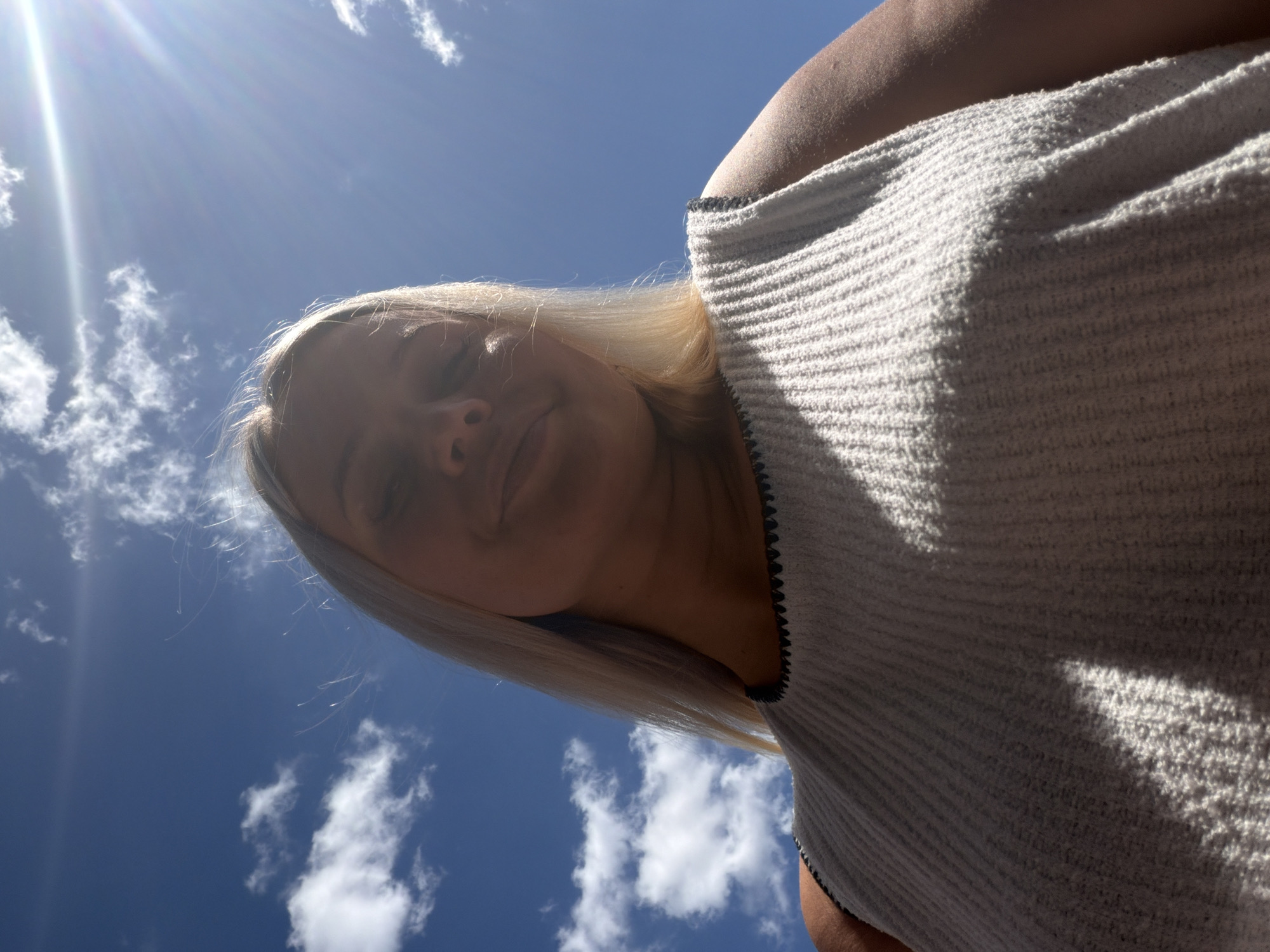 A low-angle shot captures a person with blonde hair against a bright blue sky dotted with white clouds. The sun is visible in the upper left corner, casting rays and creating a lens flare. The person is wearing a light-colored, ribbed top with a dark trim along the neckline. Their face is partially obscured by their hair and the angle of the shot, but they appear to be looking downwards with a relaxed or content expression. The sunlight highlights their skin and hair. The overall impression is one of a sunny day, possibly outdoors, with the camera positioned below the subject.