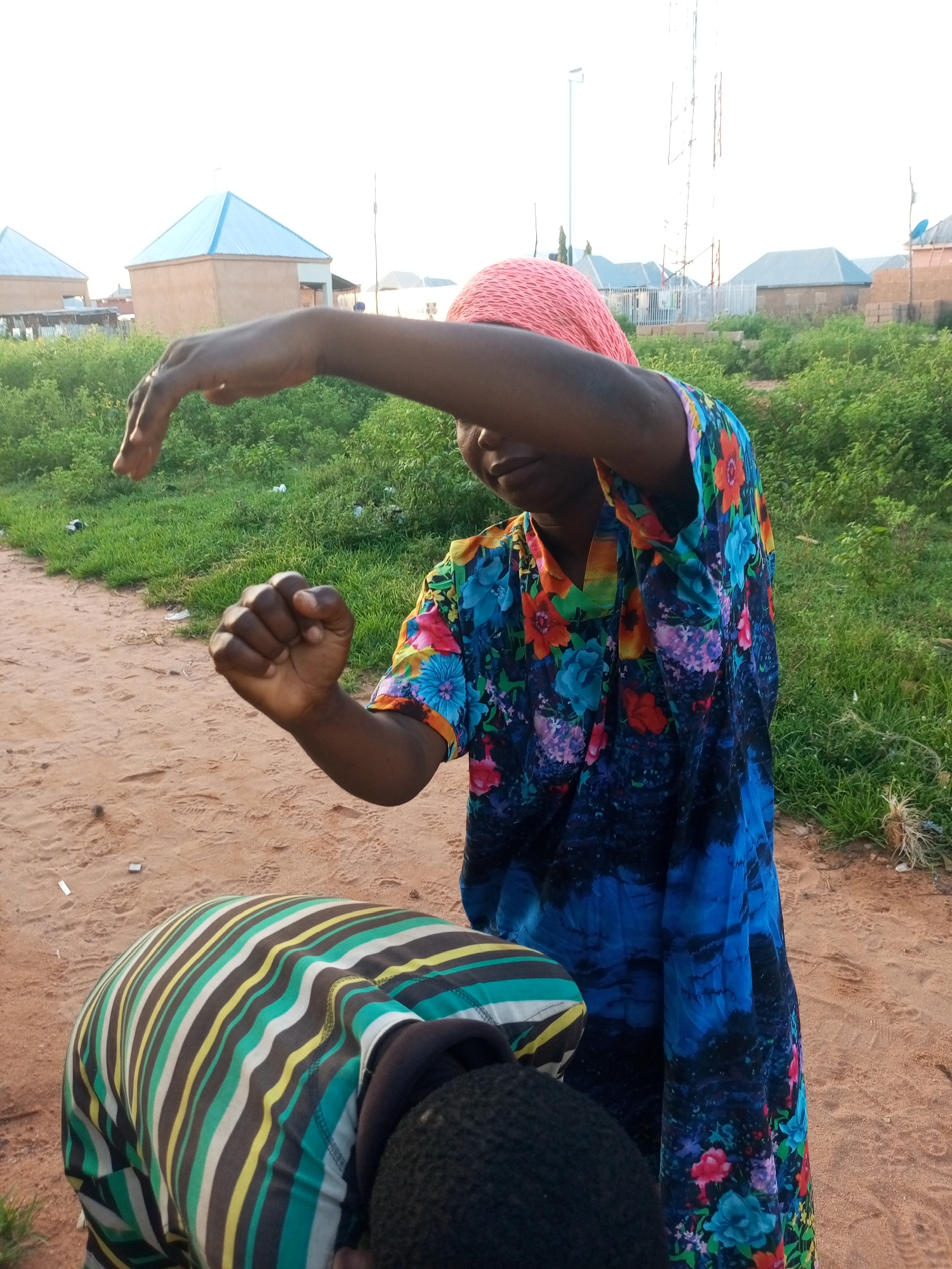 An adult woman wearing a pink head covering and a colorful floral dress stands in an outdoor environment with two other people. She is in the foreground, slightly to the right, and is extending her left arm upwards, palm facing outwards, as if signaling or playing a game. Her right arm is bent at the elbow, with her fist clenched. To her left and slightly in front of her, another person is hunched over, wearing a striped shirt. The ground is sandy and reddish-brown, with some sparse green vegetation and debris visible. In the background, there are several buildings with blue roofs and what appears to be a communication tower. The sky is bright and overcast. The location appears to be a semi-rural or developing area within the city of Tempure, Nigeria.