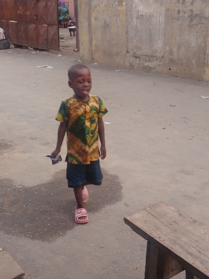 A young child is walking in a street. The child is wearing a patterned shirt with green and brown colors, denim shorts, and pink sandals. In their left hand, they hold an object. The street appears to be in an urban area, with buildings and gates visible in the background. A wooden bench is partially visible on the right side of the image. The street surface is concrete and appears to be wet in places.