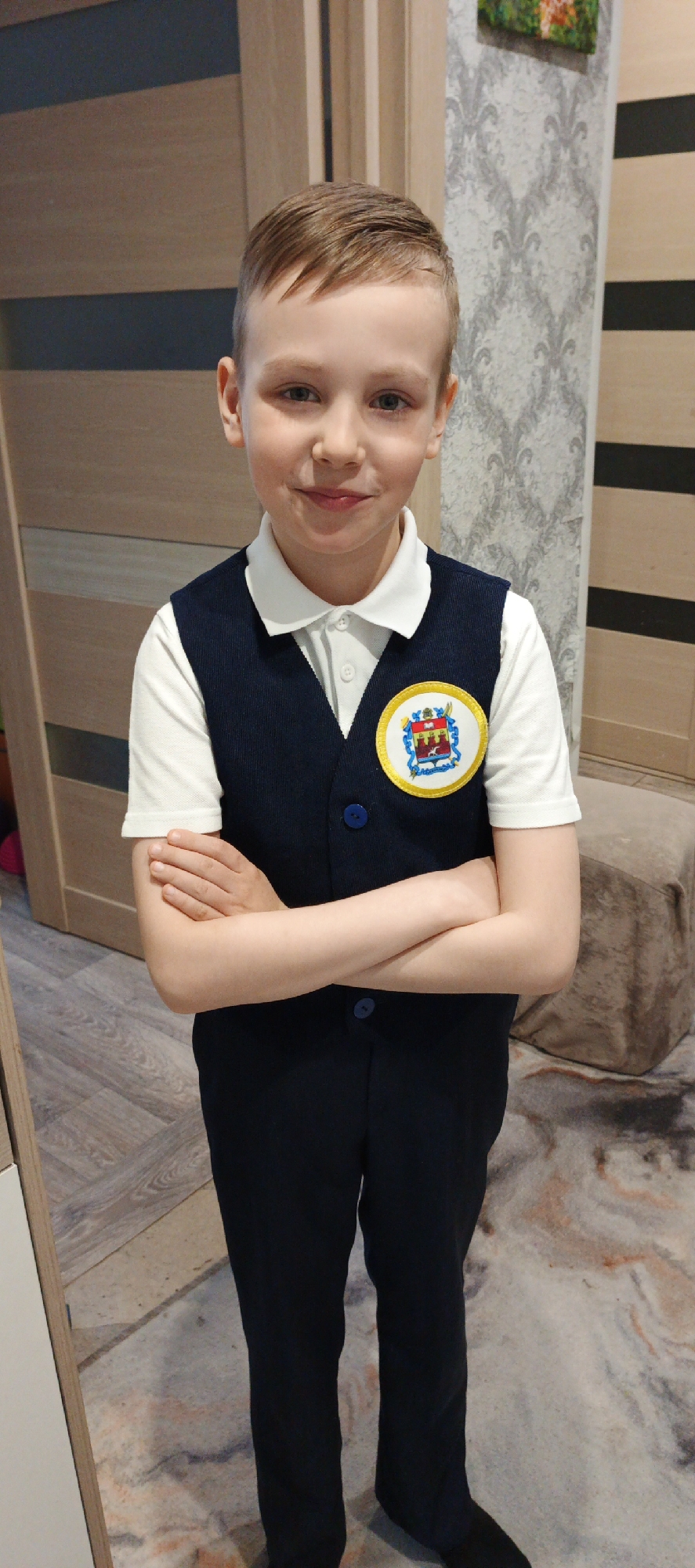 A young boy stands indoors, arms crossed, wearing a white polo shirt, a dark blue vest with matching dark blue pants, and black shoes. A circular patch with a coat of arms is affixed to the upper right side of his vest. The background includes a light brown wooden door with dark accents to the left, and a light gray patterned wallpaper with dark gray horizontal stripes to the right. A light brown cushioned cube-shaped stool is visible behind him on the right. The flooring has a patterned surface with beige and light gray tones. This is likely a school uniform with the coat of arms for the city of Chaikovsky, Russia.