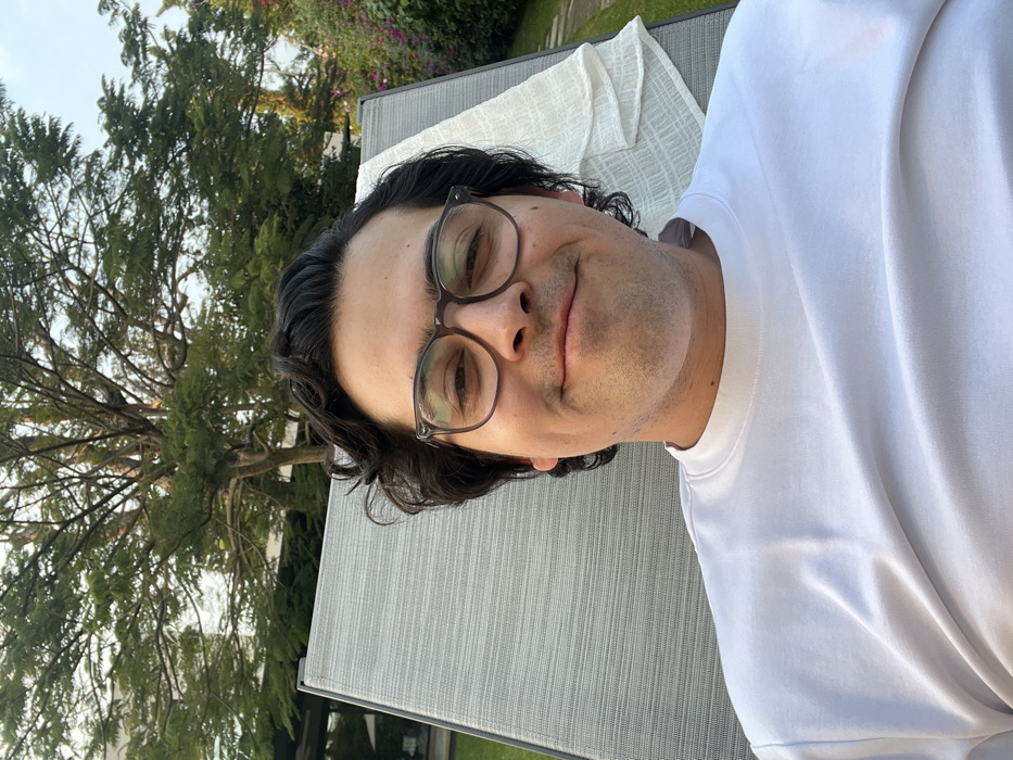 A man with dark hair and glasses is reclining on a light gray chaise lounge. He is wearing a white t-shirt. A lush green tree is visible in the background to the left. The photo appears to be taken outdoors in a garden or yard setting. The lighting is soft and natural, suggesting it is daytime.