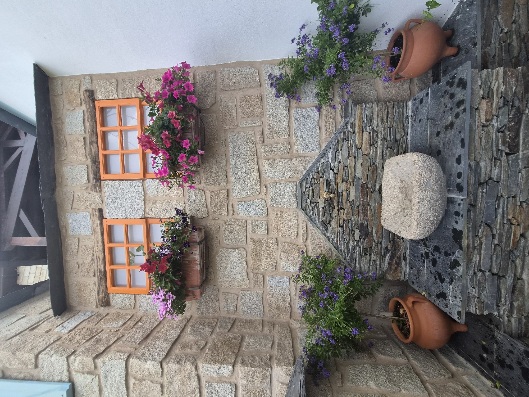 The image shows a rustic corner of a stone building in Seia, Portugal. Two small windows with orange frames are set into the stone wall, each adorned with a planter box overflowing with colorful flowers. Below the windows, a small stone fountain with a single faucet is built into the wall. A carved stone basin sits below the faucet, designed to catch water. The area around the fountain is paved with dark, irregular stones, some of which appear wet. Two potted plants with small purple flowers flank the fountain. The building's exterior is constructed from rough-hewn, multi-colored stones of various sizes, with visible mortar between them. The scene is set outdoors, with evidence of a natural, earthy aesthetic.