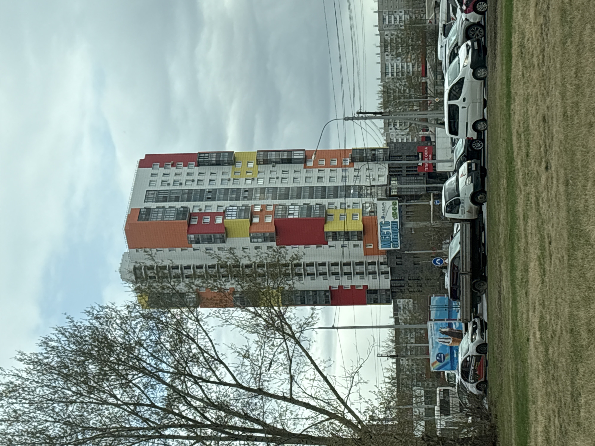 The image captures a view of Krasnoyarsk, Russia, showcasing a tall, modern apartment building with a distinctive colorful facade of red, orange, and yellow panels. The building dominates the frame and stands out against the cloudy sky. The presence of trees with bare branches suggests it may be either early spring or late autumn. A line of white vans is parked along the roadside, bordering a grassy area. Advertising signs and other buildings can be seen in the background, indicating an urban setting. Electrical wires traverse the skyline.