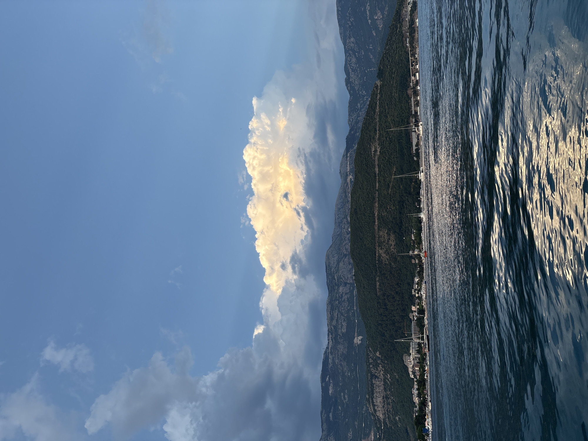 This is a landscape photograph taken from the water, looking towards a coastline with mountains in the background. The scene is oriented vertically, with the sky at the top and the water at the bottom.
The sky is a vibrant blue, with a large, fluffy white cloud illuminated by the sun, casting a warm, golden glow. Other clouds are scattered across the sky, some appearing darker and more diffused.
Along the coastline, there is a strip of buildings, suggesting a town or city. Dense green vegetation covers the lower slopes of the imposing, dark mountains that rise steeply behind the settlement. Several sailboats with tall masts are visible near the shore, indicating a maritime setting. The water in the foreground is choppy, with sunlight reflecting off its surface, creating ripples and highlights.
The lighting suggests it is either late afternoon or early morning, with the sun low in the sky and casting long shadows. The weather appears pleasant, with clear skies in some areas and dramatic cloud formations in others. The overall mood is serene and picturesque, capturing the beauty of a coastal landscape.
No visible text is present in the image. The location is identified as Budva, Montenegro.