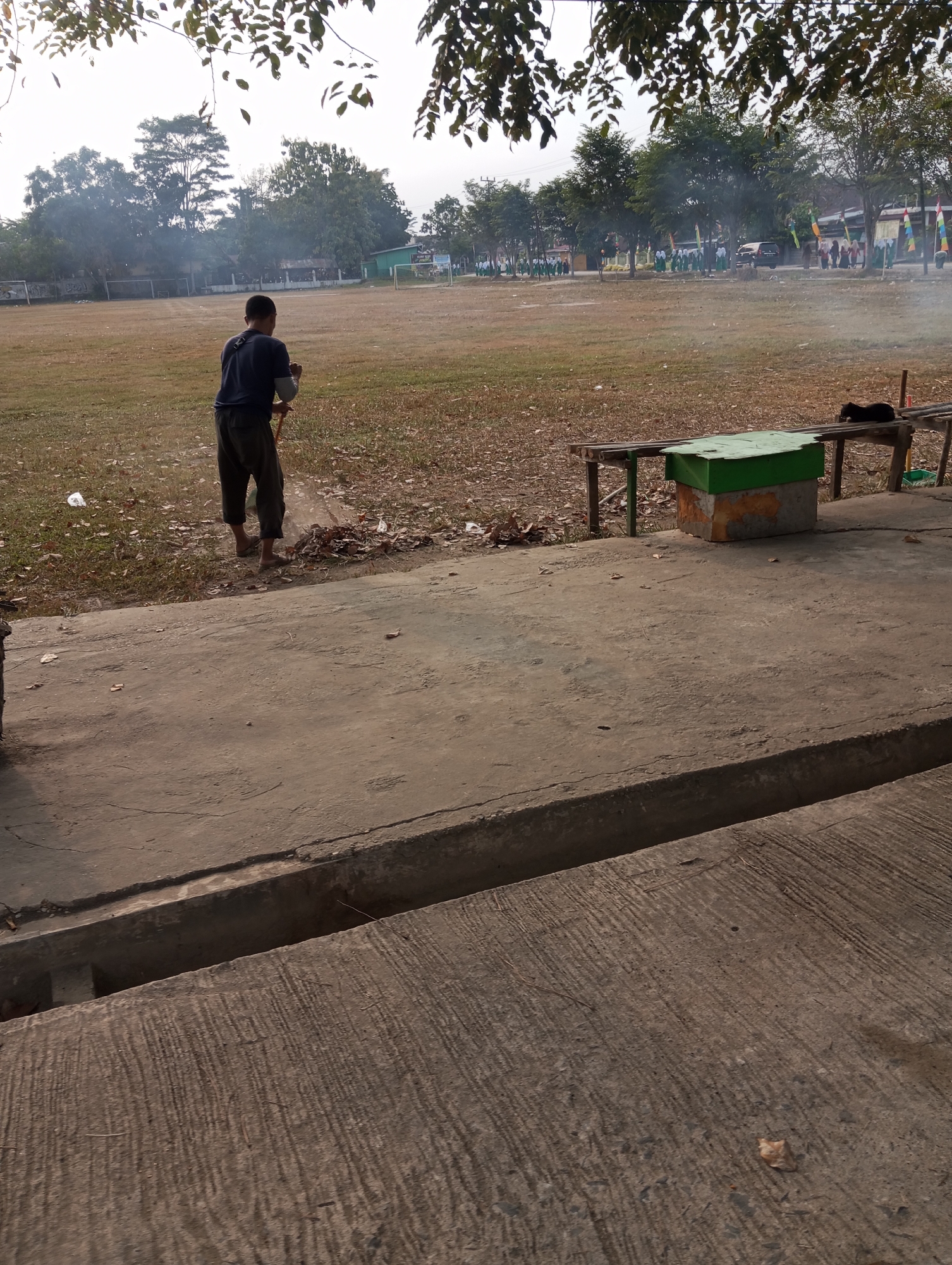 A person is sweeping dry leaves with a broom on the edge of a concrete path. Behind the person is a large, dry, grassy field. In the background, there are trees and some buildings. To the right of the person and field, there is a wooden bench with a green box on it, and a black cat is lying on the bench. The scene appears to be outdoors in a somewhat undeveloped area. The overall lighting suggests it is daytime, possibly late afternoon. The location is Pematang Reba, Indonesia.
