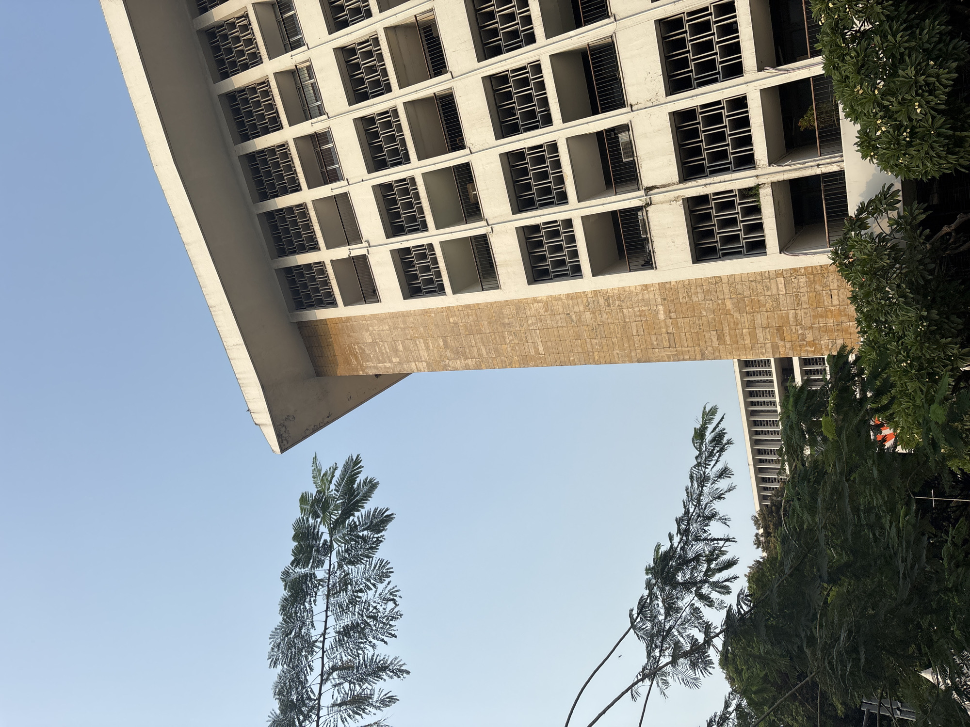 This image depicts the exterior of a building with a distinct architectural style, characterized by its repetitive window patterns and balconies, located in West Jakarta, Indonesia. The building appears to be constructed with white concrete and beige stone accents. Lush green trees partially obscure the lower sections of the building, suggesting a tropical climate. The sky above is a clear, pale blue, indicating daylight hours. The perspective of the photograph is from a low angle, looking upwards towards the building and the sky. The overall impression is one of an urban environment with significant green spaces.