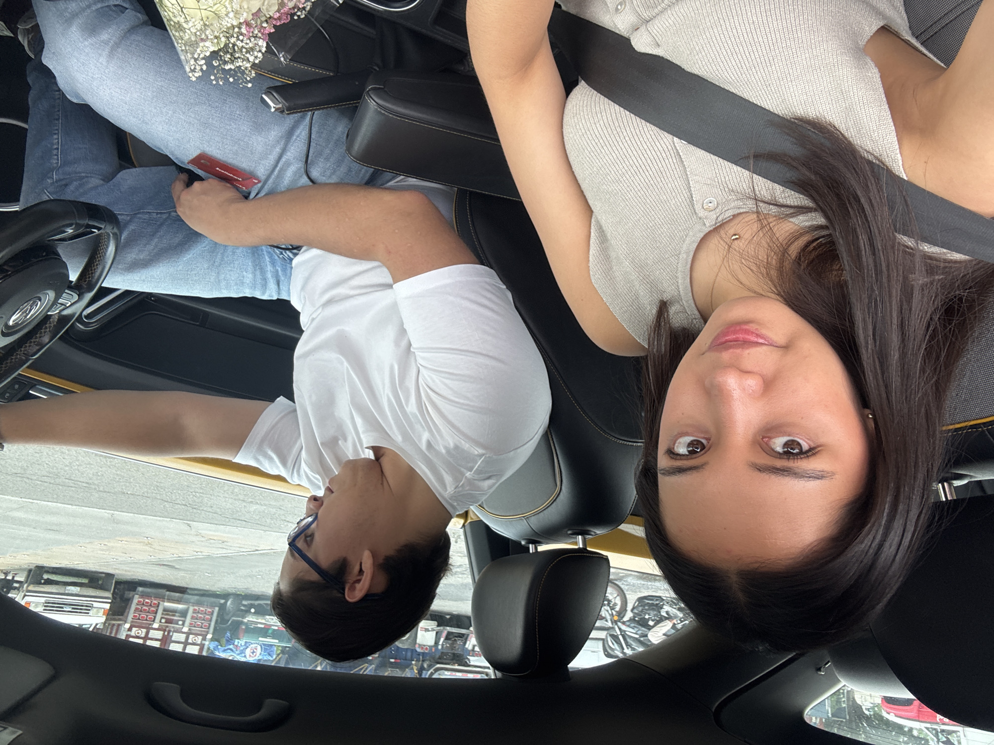 A man and a woman are in the front seats of a car. The woman is in the right seat, wearing a light-colored, button-down shirt with a seatbelt across her chest, and has dark hair. The man is in the left seat, wearing a white t-shirt and blue-framed glasses, and is reaching into his jeans pocket. There is a flower bouquet on his lap. A "Scotiabank" credit card is visible in his hand. The car is parked on a street in San Juan Tejaluca, Mexico, as indicated by the Spanish text on the "Scotiabank" card. The scene outside the car window shows buildings and vehicles.