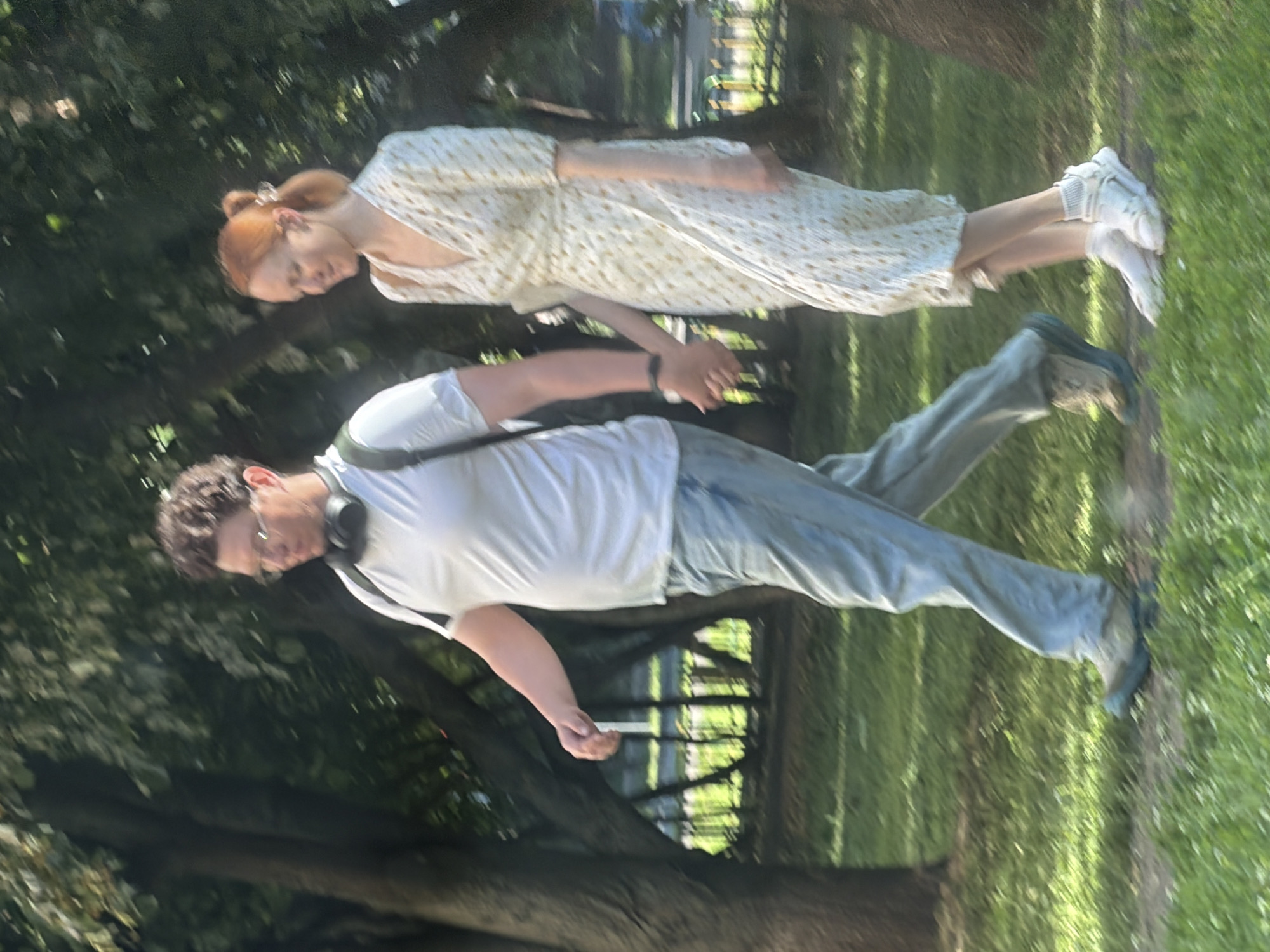 The image shows a man and a woman walking hand-in-hand on a path in a park. The man is wearing a white t-shirt, blue jeans, and sneakers. He has headphones around his neck and glasses. The woman is wearing a white dress with a floral pattern and white sandals with socks. They are walking from left to right in the frame. The background consists of trees, grass, and a fence. The lighting suggests it is daytime. The image is slightly blurred due to motion. It is not possible to determine the specific location within Moscow, Russia, from the image alone.