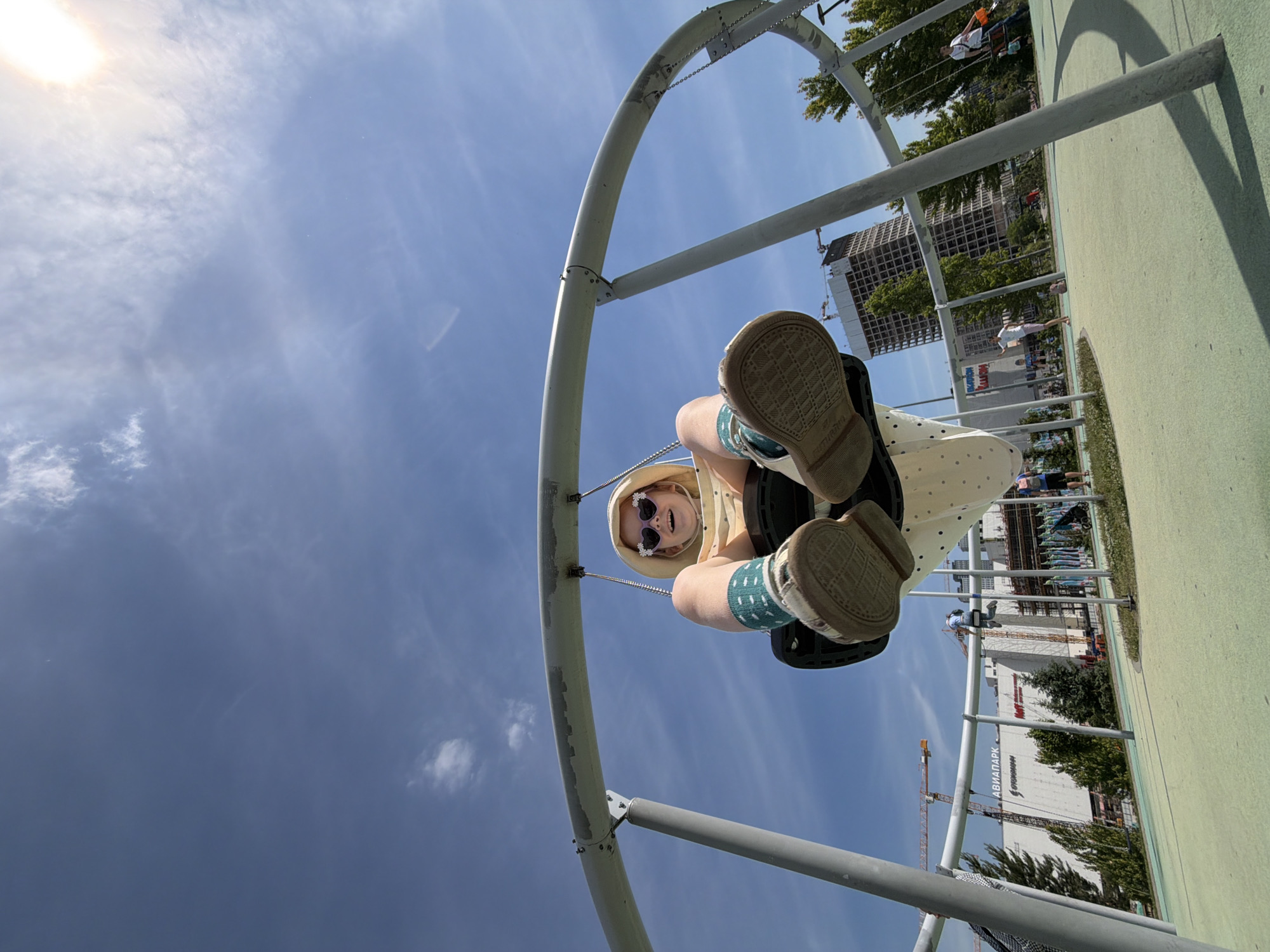 A low-angle, wide-angle shot captures a child on a swing at an outdoor playground under a bright blue sky with scattered white clouds and a visible sun flare in the upper right. The child, wearing a light-colored hooded garment, heart-shaped sunglasses, green-patterned socks, and light-colored shoes, is seen from below, elevated in the swing's upward motion, smiling with an open mouth. The swing apparatus is a large, grey metal structure featuring a curved overhead bar and stout vertical supports, with the swing seat having a black top and a white, polka-dotted bottom. The ground surface is a light green, uniform material, likely rubberized. In the background, a large white building is visible, prominently displaying "АВИАПАРК" (AVIAPARK) in red letters, accompanied by multiple construction cranes. Further to the right, a tall, multi-story building with an exposed structural grid is under construction or renovation, surrounded by green trees. Additional people are present throughout the background, utilizing other playground equipment or walking. The scene is located in the city of Khimki, Russia.