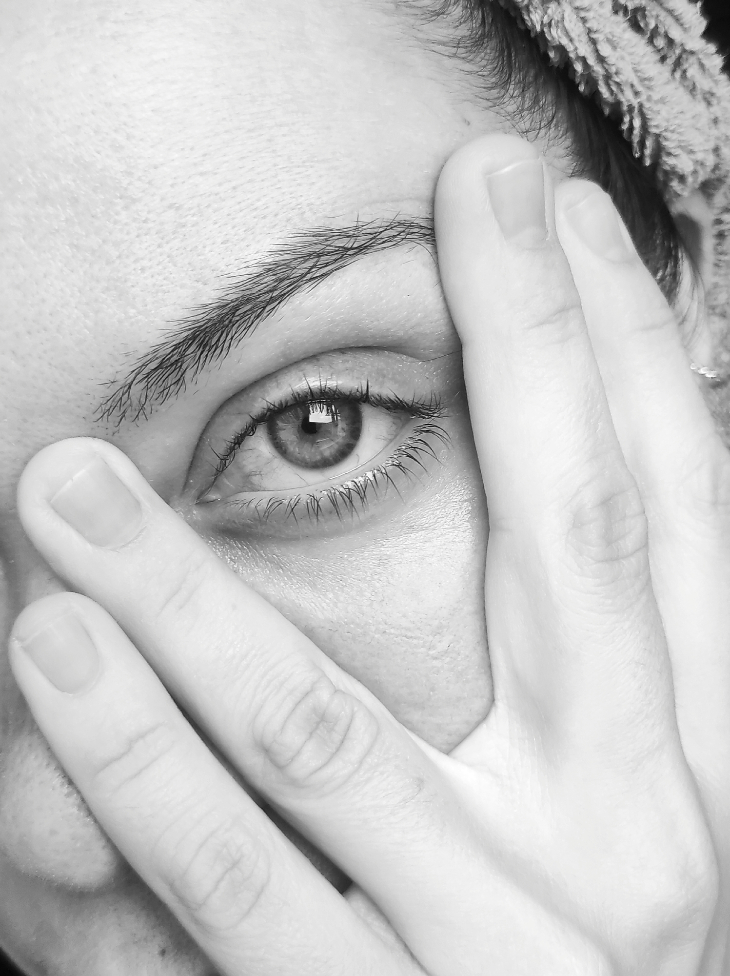 A close-up, black and white photograph focuses on a person's eye, partially obscured by their hand. The subject's left eye is visible, with a detailed iris and eyelashes. The hand is positioned in front of the face, with fingers covering part of the cheek and forehead. The lighting highlights the textures of the skin and the reflections in the eye. The background appears to be a textured surface, possibly fabric, and the edge of a towel or similar item is visible at the top of the frame. The image does not contain any elements that would allow for the identification of a specific location in Chaikovsky, Russia.