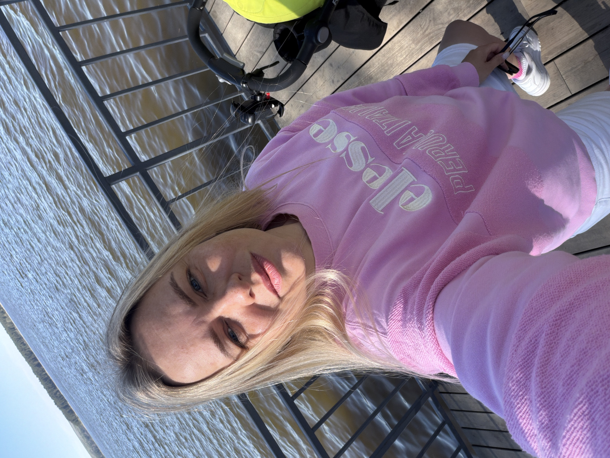 A woman is taking a selfie while sitting on a wooden deck overlooking a body of water. She has blonde hair and blue eyes, wearing a pink sweatshirt with white lettering, white shorts, and white sneakers. She is holding a pair of sunglasses in her hand. Next to her is a stroller with a green canopy. The deck has metal railings. The body of water has waves reflecting the sunlight. The city of Votkinsk, Russia is not discernable in the image.