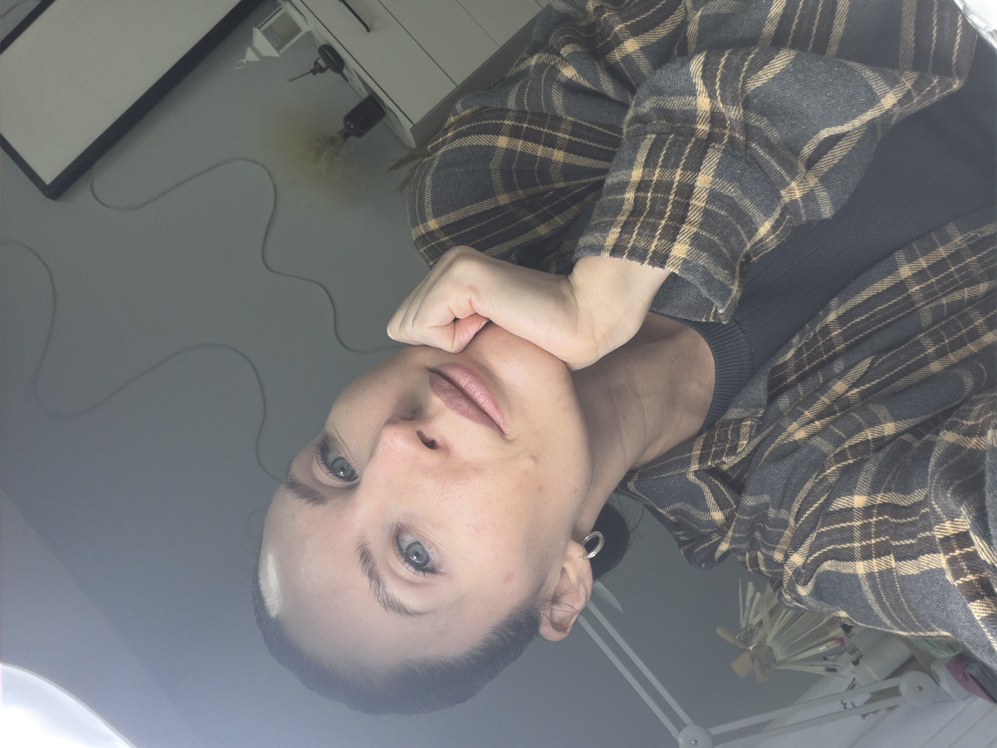 A woman with light skin and gray-blue eyes is looking upwards at the camera, resting her chin on her hand. She is wearing a dark gray sweater and a gray and yellow plaid flannel shirt. Her hair is dark and pulled back. She has small hoop earrings in her ears. The background includes a white wall, a white cabinet with what appears to be art supplies on top, and a lamp. The scene is likely indoors. The location and city cannot be determined from the image.