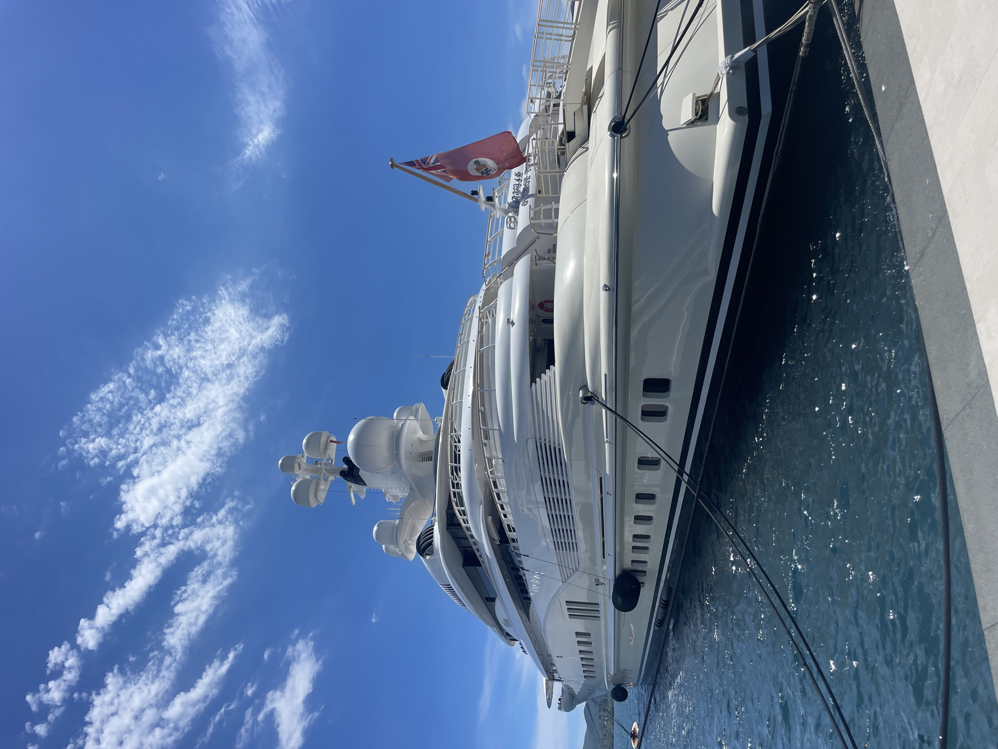 A large white luxury yacht named "Bellamus" is docked in the city of Tivat, Montenegro, on a sunny day with a blue sky and scattered white clouds. The yacht is prominently featured, taking up most of the frame. Its sleek, multi-deck structure is visible, with numerous windows lining its hull. The yacht is secured to a dock by thick black ropes. Two dark fenders are attached to the side of the yacht to prevent damage. A red flag with a crest and a Union Jack in the canton is flying from a mast on the yacht. The water around the yacht is a deep blue and shows some ripple effects from the sun. In the distance, a hazy green coastline is visible. The overall scene depicts a serene and opulent maritime setting.