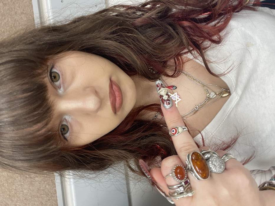 A close-up, chest-up portrait depicts an individual, likely female, with shoulder-length brown hair highlighted with reddish strands and straight bangs. Their eyes are light brown/hazel, accentuated by white eyeliner on the lower lash line. The lips are lightly colored, and the facial expression is neutral. The individual wears a white V-neck shirt.
The right hand's index finger is prominently raised and pointed towards the left. This finger features an elaborate, long nail with a silver-grey base, white intricate patterns, and a red "blood drip" design extending from the tip. A silver-toned ring with a red and blue "evil eye" motif is worn on this finger. The left hand, partially visible, displays multiple silver-toned rings, including one with a large oval amber-colored stone on the middle finger and another with a round orange stone on the index finger.
Around the neck, several layered silver-toned necklaces are visible. Charms include a small pink Star of David, a heart-shaped locket, and a red bead. The background is an indistinct indoor setting, featuring a light-colored, possibly textured wall on the left and a lighter, smoother surface on the right, with a horizontal light-colored rail present behind the head on the left. The specific location within Raanana, Israel, cannot be determined from the visual information provided.