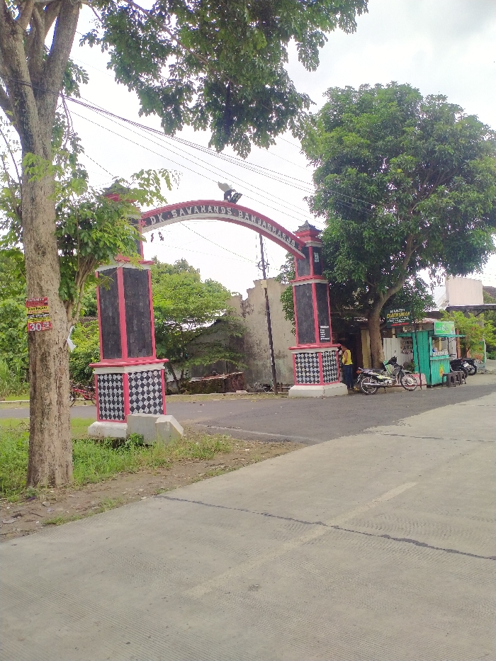The image depicts a daytime scene at the entrance to a community in Kecamatan Kebakkramat, Indonesia, marked by a decorative arched gate. The gate features two substantial pillars and an arch connecting them. The pillars are painted with red and black accents, and their bases exhibit a distinct black and white checkerboard pattern. Text on the arch clearly reads "DK. SAWAHANDS BANJARHARJO", with a light fixture mounted above it.
On the left side of the frame, a large tree trunk and its foliage partially obscure the gate. A red and white sign is attached to the tree, displaying the text "KOPI KAKI" and the number "30". To the right of the gate, another large, leafy tree stands beside a small, light blue/green street stall or shop. A person wearing a yellow shirt is visible standing near the right pillar of the gate, in proximity to the stall. Several motorcycles are parked in front of the stall, and a bicycle is partially visible near the left pillar.
The ground in the foreground is a concrete road, which changes to an asphalt surface beyond the gate. The sky appears overcast, indicating a cloudy day. In the background, behind the gate, more trees and some buildings are visible. Power lines traverse the sky above the gate. The overall scene suggests a local community entrance, possibly a casual gathering point or business area, characteristic of a rural or semi-rural setting.