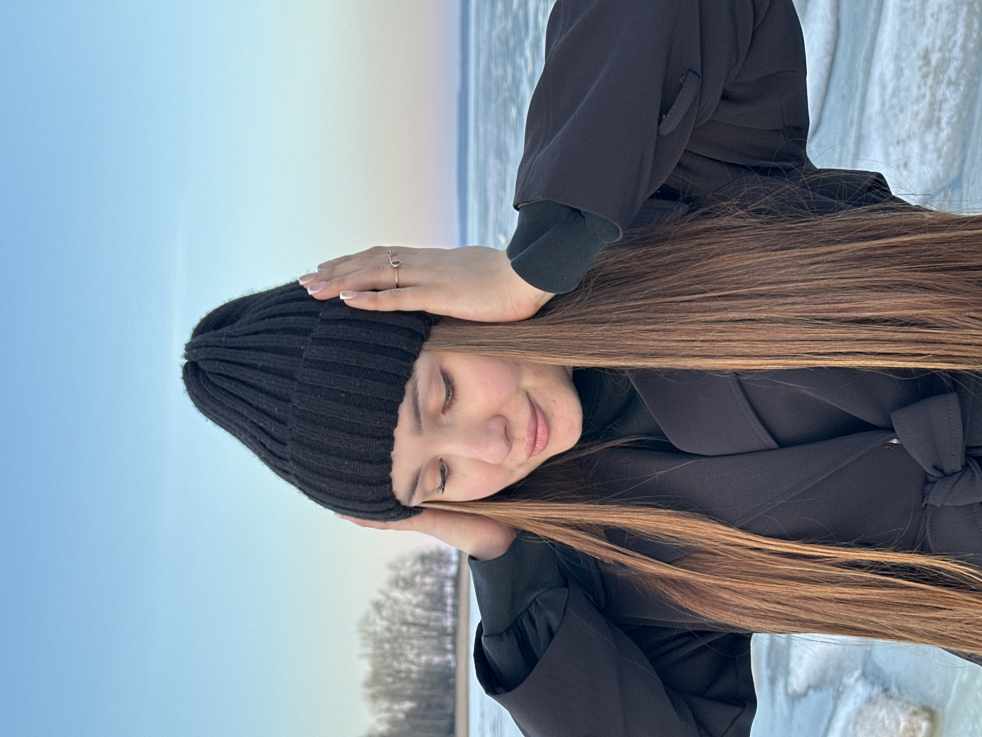 A young woman stands outdoors in Chaikovsky, Russia. She wears a black knitted hat and a dark jacket. Her long, brown hair is straight and reaches past her shoulders. She rests her head on her hand, appearing thoughtful or serene. The background shows a landscape with a body of water, possibly a frozen river or lake, and a pale sky. Bare trees are visible on the horizon, suggesting it is winter or early spring. The ground is covered in snow and ice. The lighting suggests it is daytime, but the overall tone is subdued.