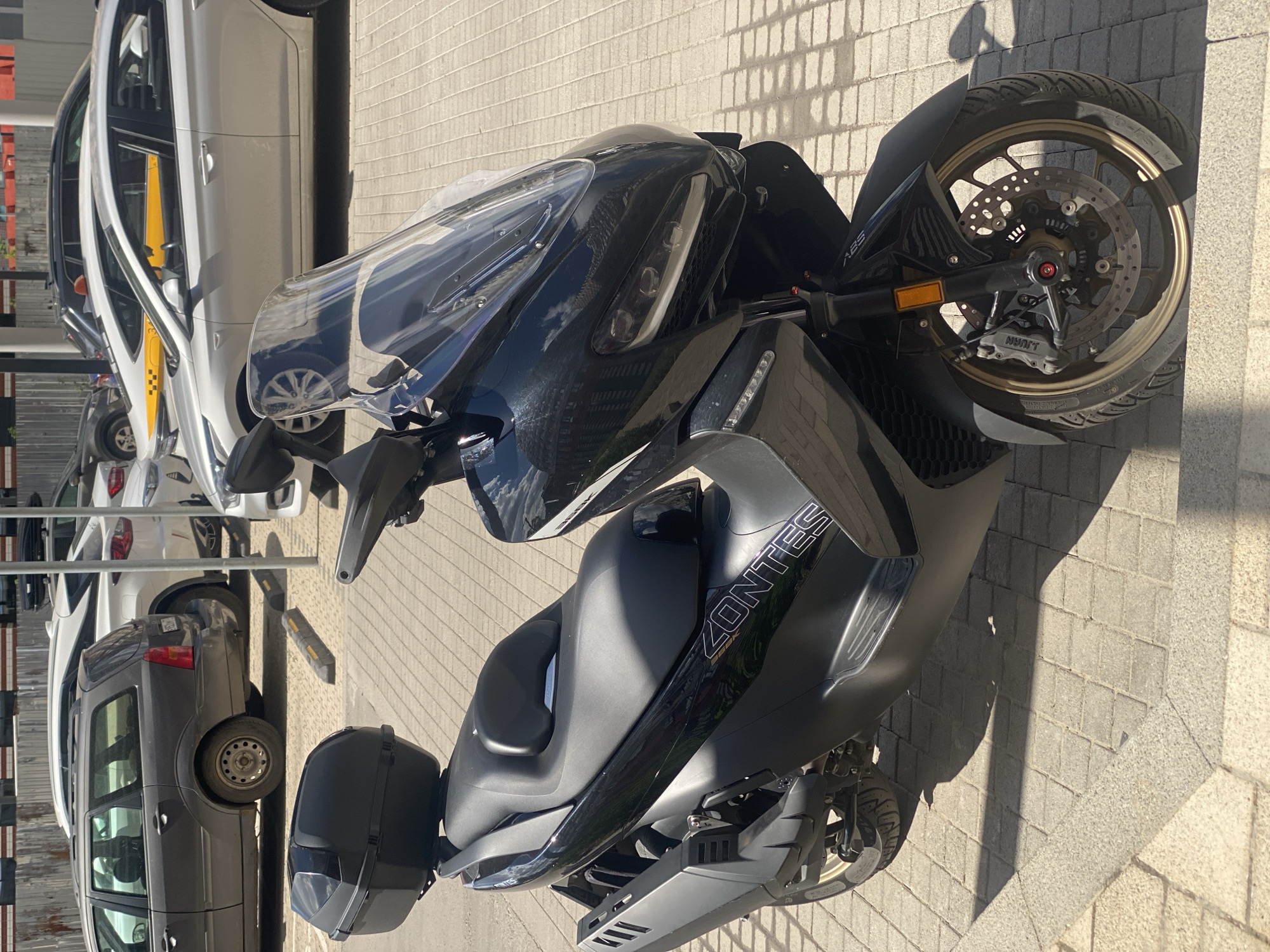 The image shows a black scooter parked on a sidewalk made of light gray paving stones. The scooter is a Zontes model, with "ZONTES" written in white lettering on its side. The scooter has a large clear windshield and black handlebars. The front wheel has an ABS system. To the left of the scooter, there are several parked cars, including a white taxi with a yellow stripe and black checkered pattern. The cars are parked in what appears to be a parking lot. The sun is casting shadows on the ground, suggesting it is daytime. The location is Kotelniki, Russia. There are no people visible in the image. The scene is still and quiet, with no specific activity taking place.