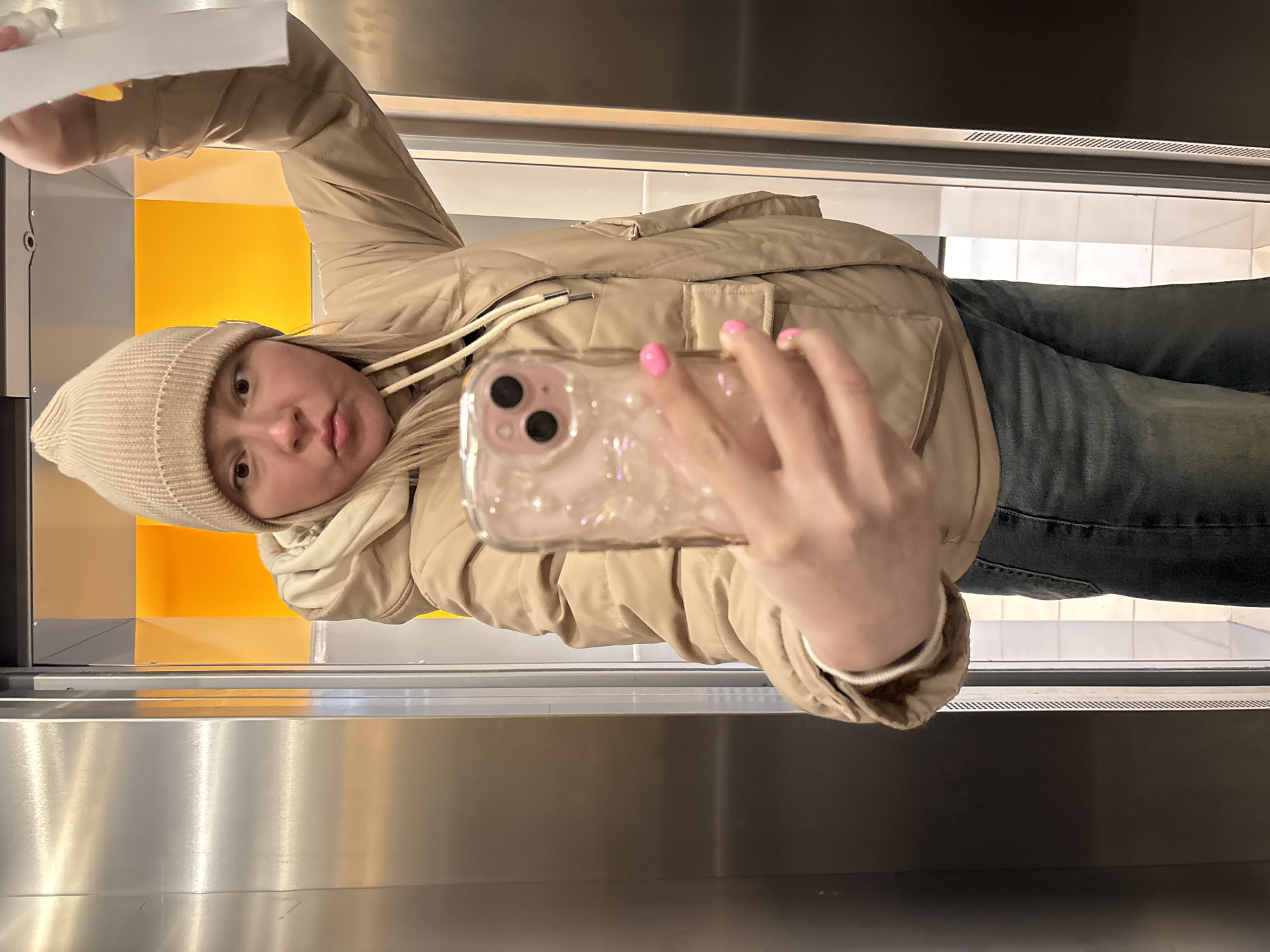 A woman takes a selfie in an elevator, possibly in Izhevsk, Russia. She wears a beige beanie, a puffer jacket, and jeans. She holds a phone with a clear, glittery case in her right hand, which has pink nail polish. The elevator has stainless steel walls and a section with yellow and white tiles. A white object is held in her left hand above her head.