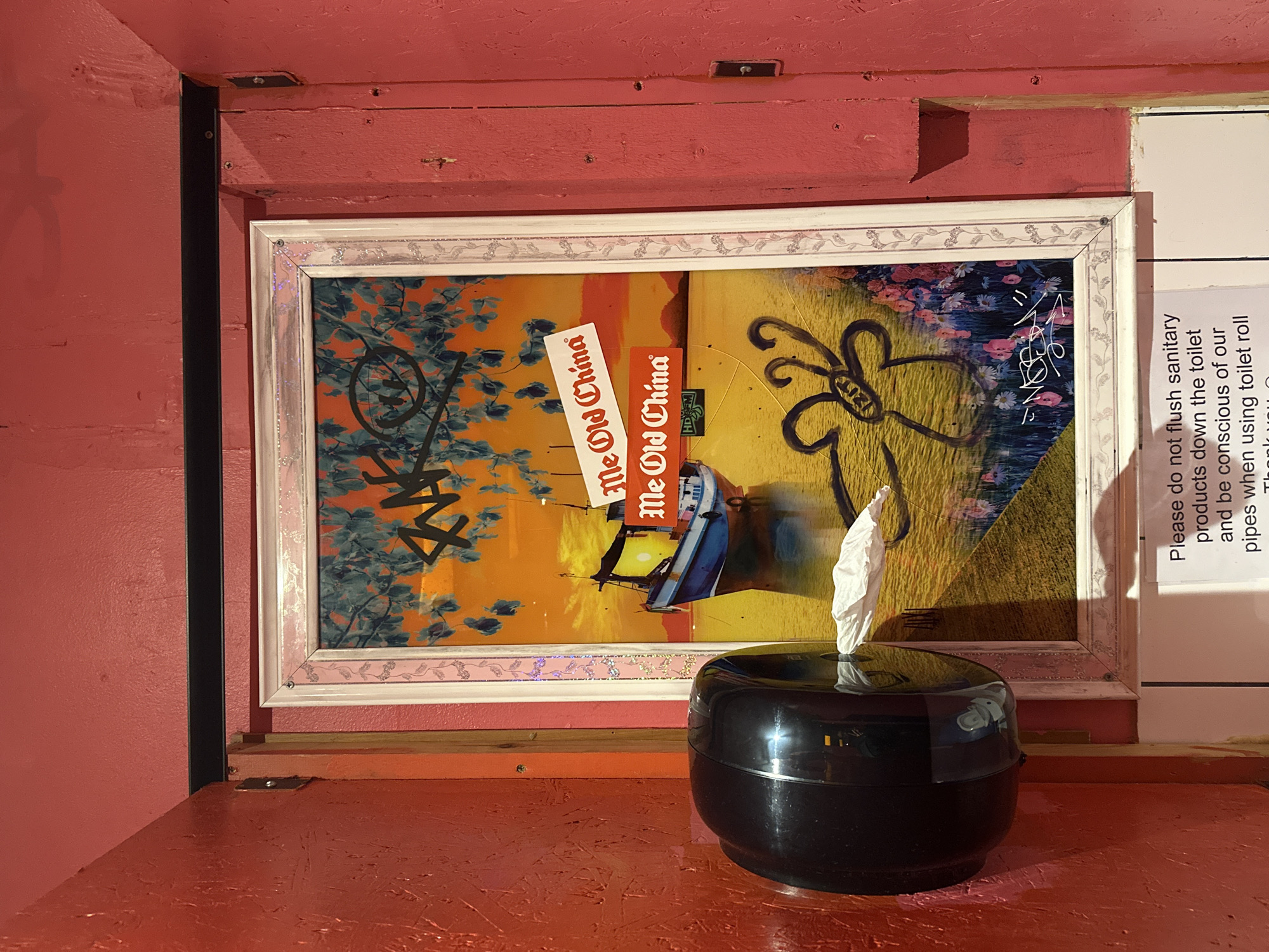 The image captures a corner of a bathroom stall in what appears to be a quirky or eclectic setting. The walls are painted in shades of red, and a framed piece of artwork hangs above a tissue dispenser on an orange countertop. The artwork is a colorful collage with images, stickers, and graffiti, and the frame is decorated with a floral pattern. A sign instructing users not to flush sanitary products down the toilet is affixed to the wall to the right of the artwork. The overall impression is of a unique and possibly repurposed space. The artwork contains "Me Old China" stickers, which may indicate a location in London, UK.