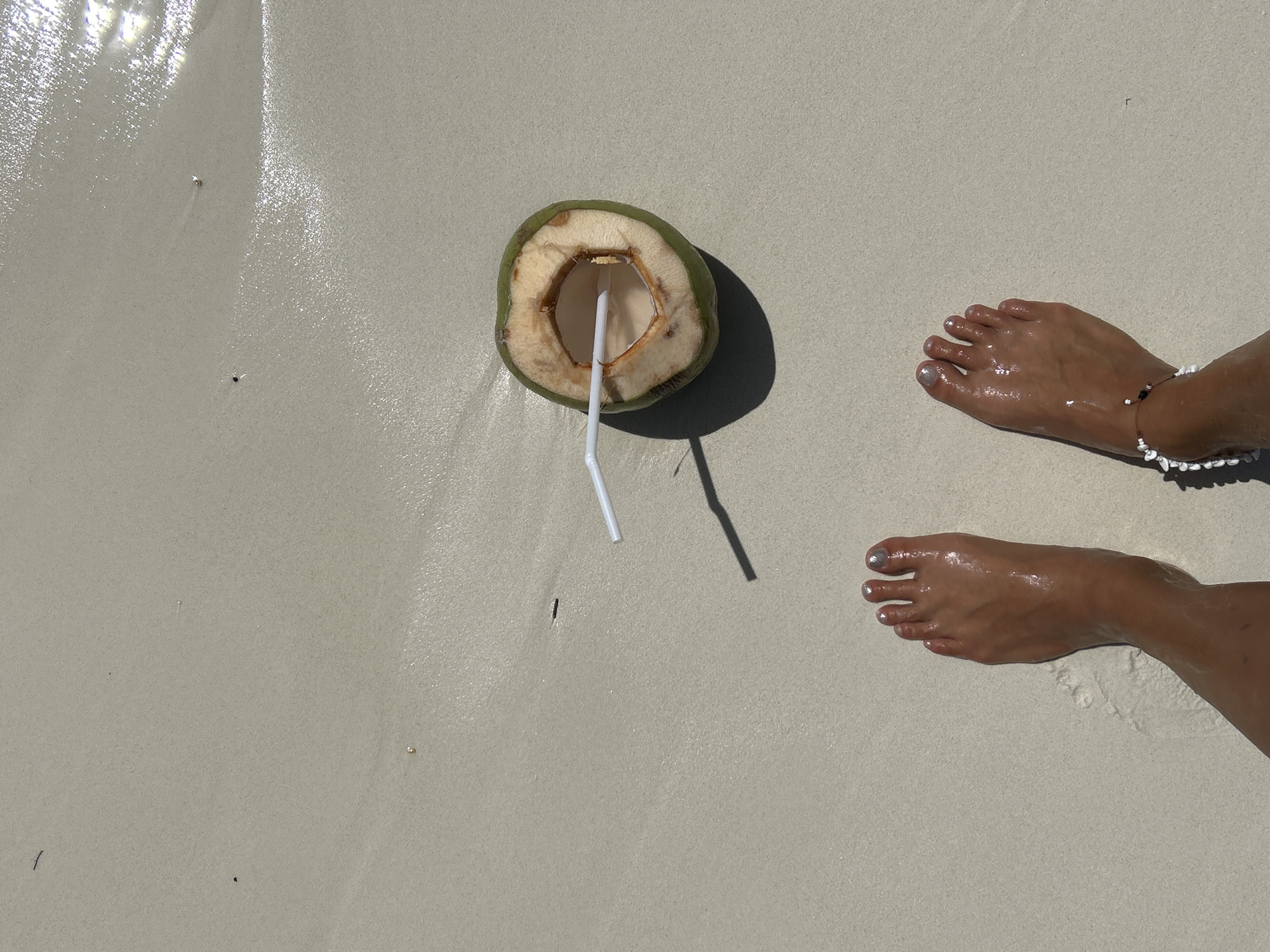 The image shows a top-down view of two wet feet on a sandy beach. A halved coconut with a straw is placed on the sand between the feet. The coconut appears to be filled with liquid. The sand is light in color and wet, with some small shells or debris visible. The water's reflection can be seen in the upper right corner of the image, indicating a shallow water line. The overall scene is consistent with a tropical beach environment in Ao Nang, Thailand.