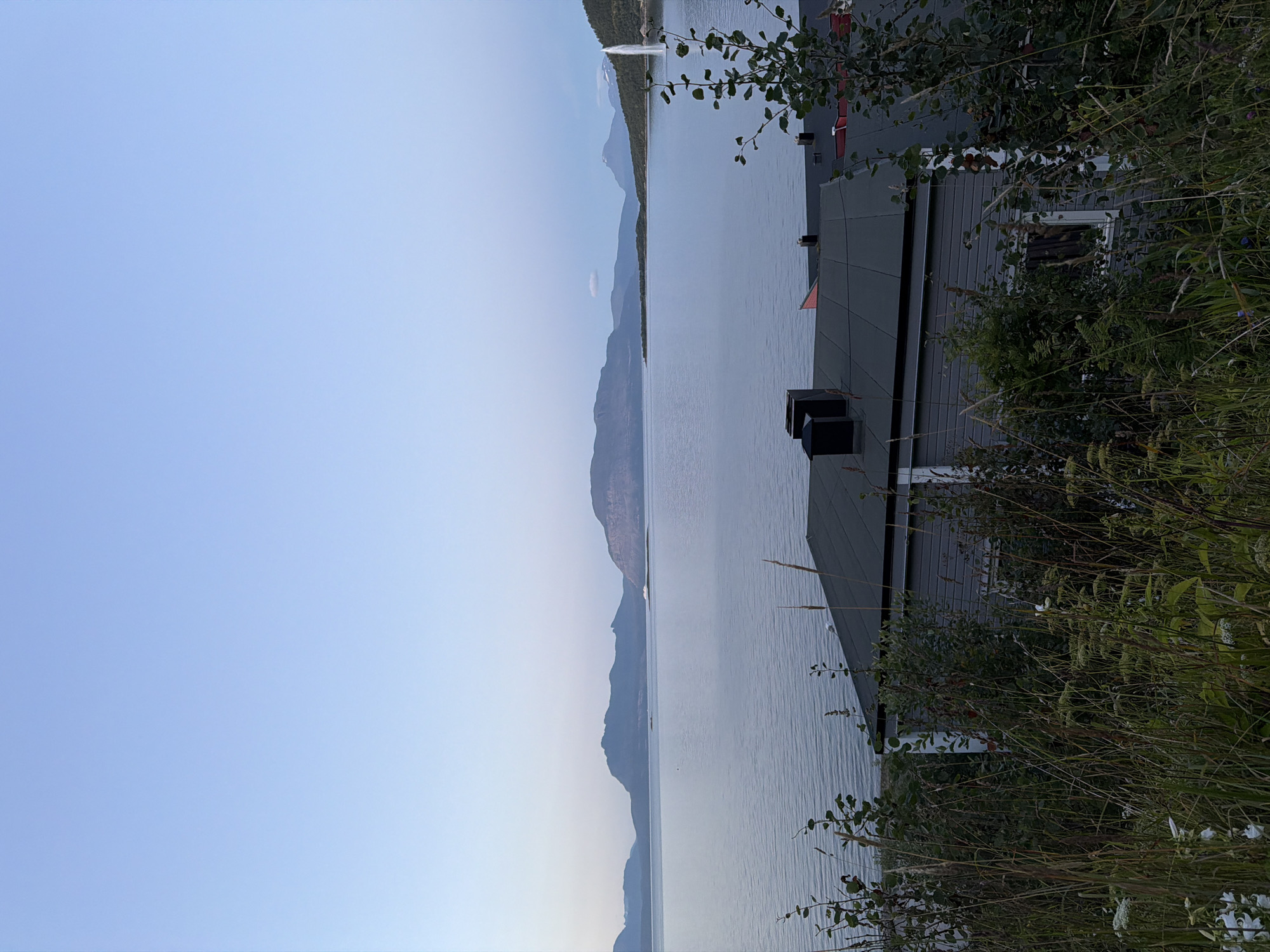 The image depicts a tranquil natural scene with a body of water, mountains in the distance, and a foreground with vegetation and buildings. The sky is a soft gradient of blue, transitioning to lighter hues near the horizon. In the middle ground, a vast expanse of water stretches out, reflecting the muted colors of the sky and the surrounding landscape. Distant mountains, characterized by their rugged outlines and varied elevations, form a majestic backdrop. Some of these mountains appear to be covered in sparse vegetation or rock.
In the foreground, tall grasses and other wild plants create a natural barrier, obscuring some of the lower parts of the scene. Partially visible behind this vegetation are the roofs of several buildings, appearing to be houses. These structures have dark, sloped roofs, with some featuring chimneys or ventilation pipes. One building is primarily gray with white trim around windows, suggesting a residential or possibly commercial property. Red roofs are also visible on other structures.
To the far right of the image, a tall, thin plume of white water is erupting upwards from the surface of the lake, indicating a geyser or a fountain. The overall impression is one of a serene, perhaps remote, setting, likely in a northern climate given the subtle lighting and the mountainous terrain.
Considering the provided location of Harstad, Norway, this scene could be a view of the Vågsfjorden, a fjord that runs through the city and is surrounded by characteristic Norwegian mountains. The presence of houses and the natural landscape strongly suggest a coastal or fjord-side setting typical of this region. The tall water plume might be a man-made feature or a natural phenomenon, but its prominence suggests it is a point of interest in the scene.