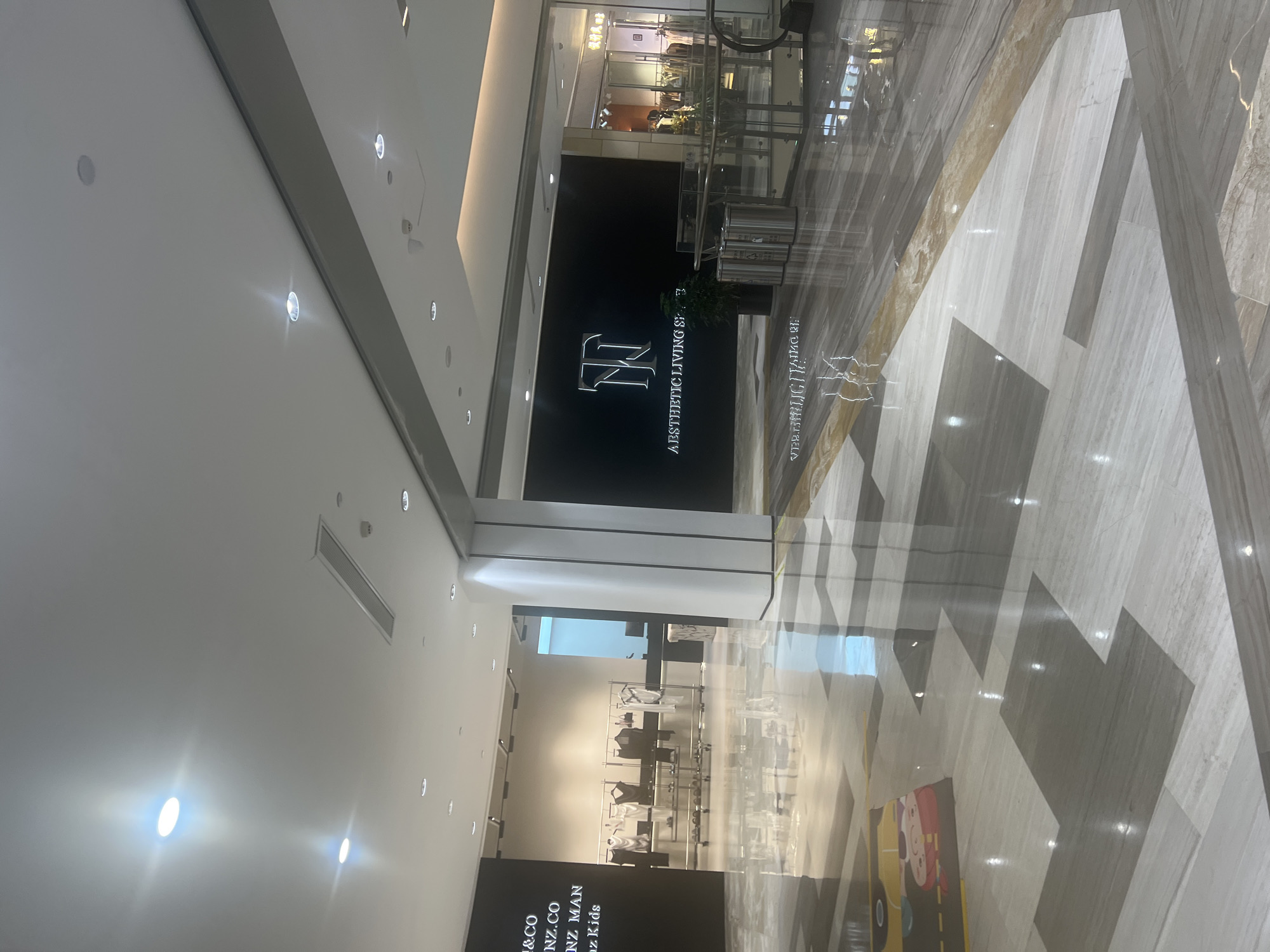 The image depicts an interior view of a modern shopping mall in Liandu District, China. The scene is well-lit with a combination of recessed ceiling lights and ambient mall lighting. The flooring is a polished marble or similar stone, laid in a geometric pattern of light and dark grey and white tiles, with a golden accent stripe running through it. The reflective surface of the floor mirrors the lights and structures above.
On the left side of the image, a store's dark facade displays text, partially visible, including what appears to be "&CO", "NZ.CO", "NZ MAN", and "oz Kids". In front of this store, racks of clothing are displayed on a white tiled floor. The clothing items are predominantly black and white, appearing to be fashion garments.
To the right, a large, black wall features a stylized logo and the text "AESTHETIC LIVING SPACE" in white, illuminated lettering. Adjacent to this wall are three silver cylindrical trash receptacles. Further to the right, a section of the mall shows store windows with clothing displays and escalators leading upwards, suggesting multiple levels. The overall environment is clean, spacious, and designed for retail shopping. There are no visible people in the immediate foreground, though there may be some indistinct figures within the storefronts on the right.