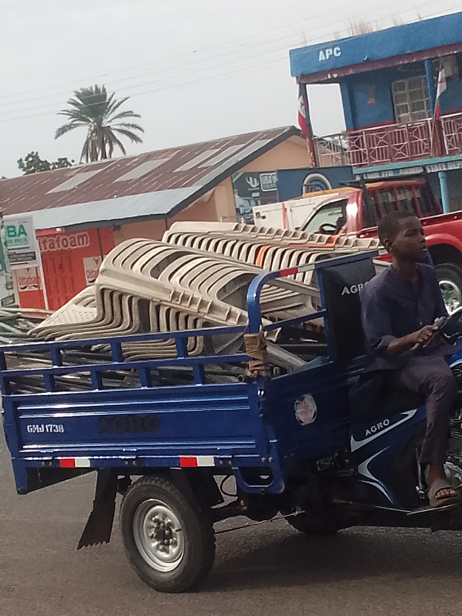 In Pantame, Nigeria, a blue three-wheeled vehicle, identified by a license plate that reads "GMJ 1738," transports a stack of beige plastic chairs. The vehicle is being driven by a young man wearing a navy blue shirt and dark pants, his right hand on the steering mechanism. Behind the vehicle, a red truck is visible, and further in the background, buildings with corrugated metal roofs are present. One building is painted blue with the letters "APC" prominently displayed on its facade, and another building features a red sign with the word "Itafoam." A palm tree stands tall against a cloudy sky.