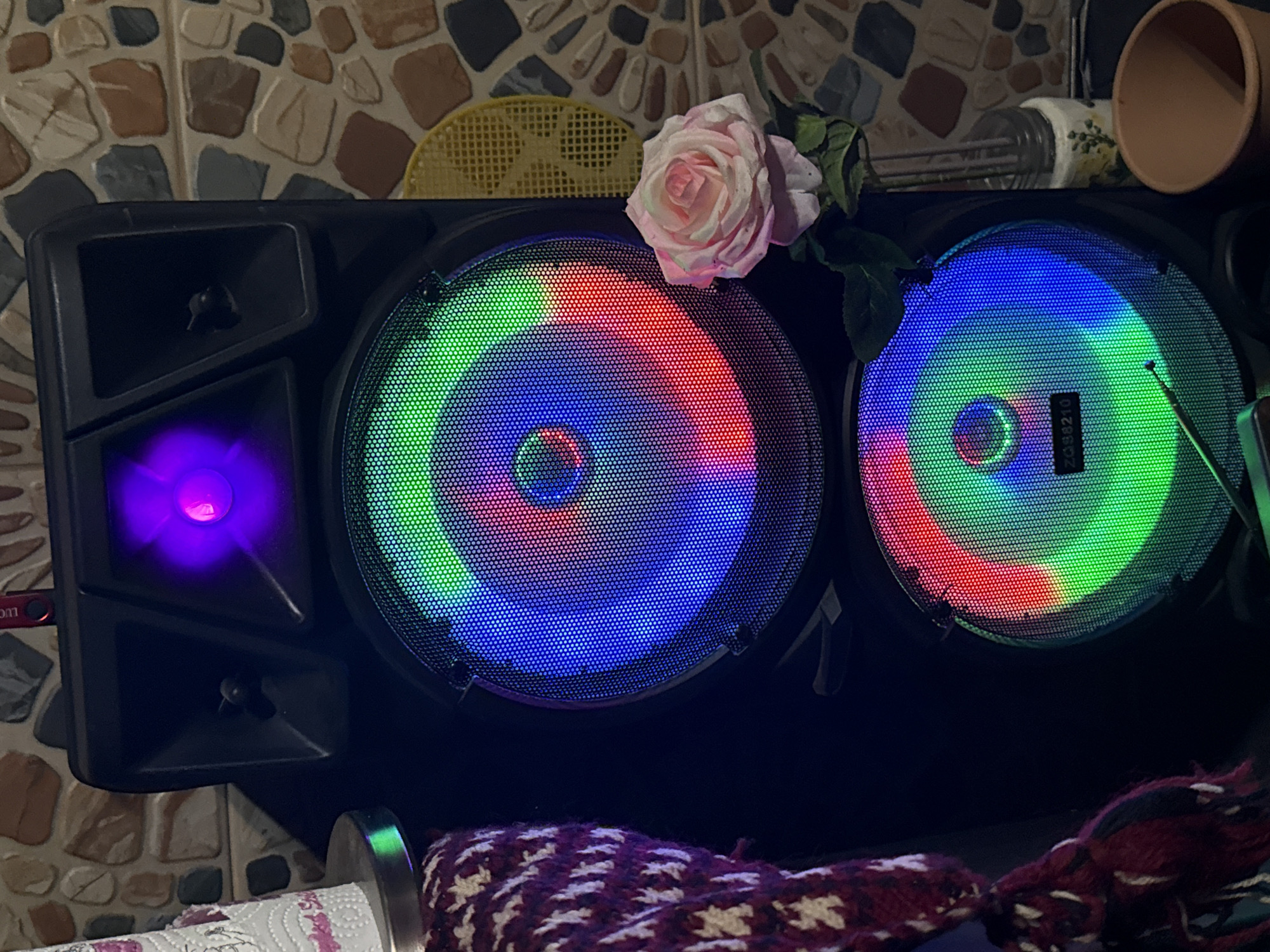 This is a close-up, eye-level photograph of a large, black, portable speaker with multi-colored LED lights. The speaker has two prominent circular grilles, each with a diameter of approximately 10 inches, displaying vibrant red, green, and blue lights. Above these, there is a smaller, triangular grille with a purple LED light. To the left of the speaker, a portion of a white paper towel roll with pink floral patterns is visible, resting on a metallic base. To the right of the speaker, a single pink and white rose with green leaves is positioned in front of a decorative jar and a terracotta pot. The background consists of a wall with a mosaic of irregularly shaped stones in various earthy tones, such as brown, beige, and grey. The lighting in the scene is dominated by the colorful LEDs of the speaker. The location of this image is the city of Sarnevo, Bulgaria.