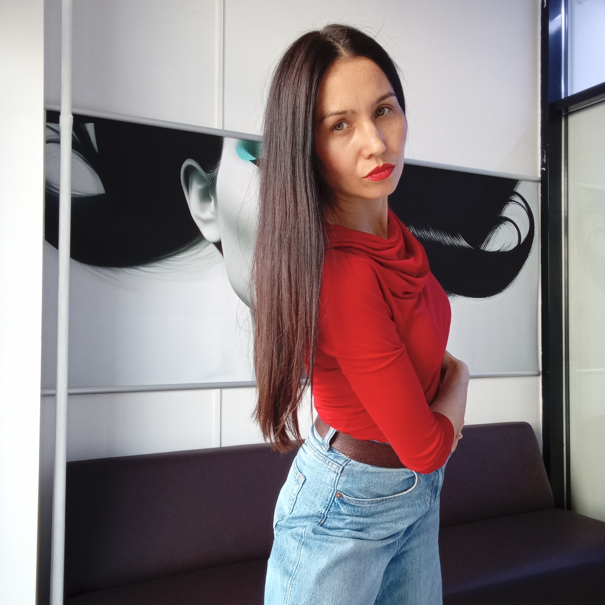 The image captures a woman posing indoors, likely within a modern office, lounge, or commercial setting in Izhevsk, Russia. The main subject, a woman with long, straight dark brown hair parted down the middle, stands with her body slightly angled to the left, looking directly at the camera. She wears a vibrant red long-sleeved top with a cowl neck, paired with light blue denim jeans and a dark brown belt. Her lips are painted with bright red lipstick, complementing her composed expression and light brown eyes.
Behind the woman, a large, stylized black and white artwork is displayed on a white wall. This artwork features a profile of a woman's head with voluminous dark hair and a visible ear, accented with a subtle turquoise hue around the eye area. Below the artwork and partially visible beneath the woman is a dark-colored sofa or bench, suggesting a seating area.
The background consists of clean white walls, possibly paneled, with a vertical white column on the left side of the frame. On the right, a dark-framed section, possibly a window or door frame, adds to the contemporary interior aesthetic. The even lighting suggests an indoor setting, likely with artificial light, creating a clear and focused portrait within the sleek environment. The woman's posture indicates she is deliberately posing for the photograph.