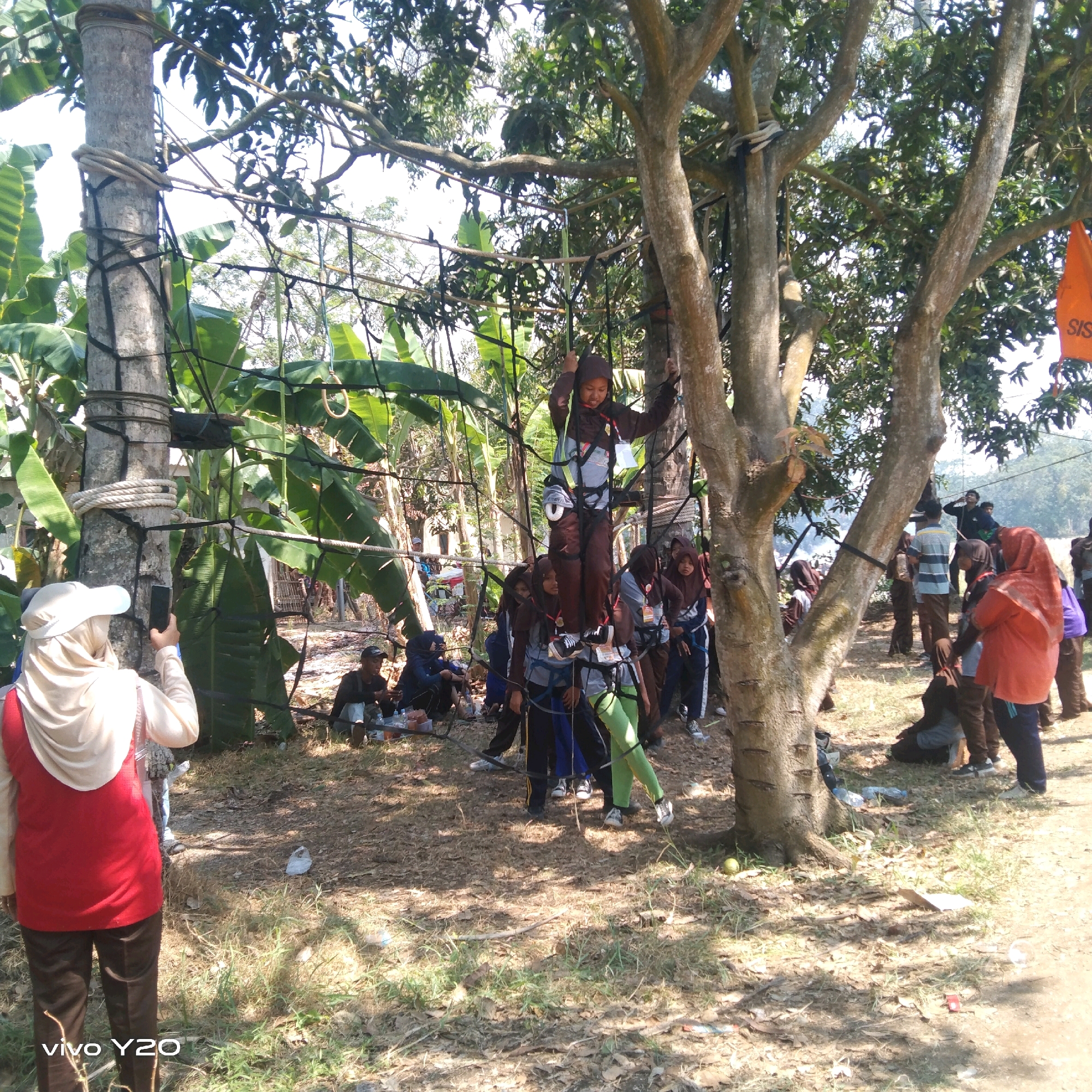 A group of young people, likely students, are engaged in an outdoor activity involving a rope obstacle course set up between trees. In the foreground, a person wearing a light-colored headscarf and a red vest is holding up a smartphone, seemingly taking a picture or video. The obstacle course consists of nets and ropes stretched between trees, with some participants actively navigating it. One individual, a young person in a brown hijab and dark clothing, is positioned high on the course, holding onto ropes. Several other young people, dressed in similar attire (hijabs and modest clothing), are visible either on the course, preparing to participate, or observing from the ground. Some are seated on the ground, while others stand. The environment appears to be a natural setting with trees, foliage, and dry ground covered with leaves and grass. Banana trees are visible in the background, suggesting a tropical or subtropical climate. An orange flag with "SIS" printed on it is attached to a tree in the upper right portion of the image. The scene is illuminated by daylight, casting shadows on the ground. The presence of the banana trees and the general appearance of the environment suggest a location in a rural or semi-rural area, potentially a park or recreational site. The overall impression is one of an organized outdoor educational or recreational event.