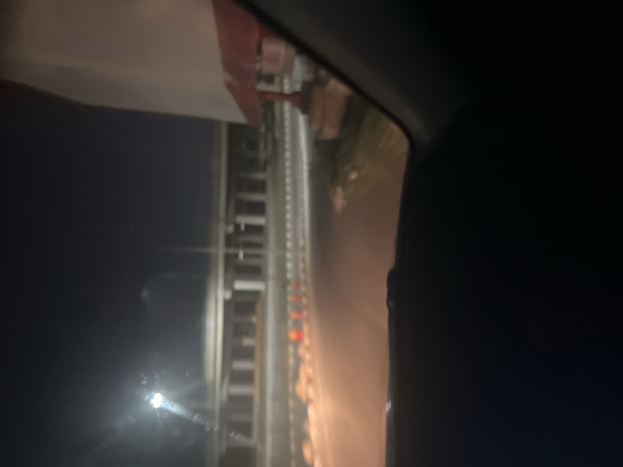 The image captures a nighttime scene in Aso, Nigeria, from the interior of a vehicle, looking out at a brightly lit structure. The primary subject is a section of what appears to be a roller coaster track or a similar amusement park ride. The track is constructed from metal supports and runs horizontally across the frame.
Along the track, a series of small, distinct lights, predominantly red, orange, and yellow, illuminate parts of the structure, suggesting decorative lighting or operational indicators for the ride. A prominent, bright light source, possibly a headlight or a streetlamp, is visible towards the left side of the frame, casting a significant glare or reflection on the vehicle's window.
The foreground and background are mostly shrouded in darkness, indicating the late hour. Parts of the vehicle's interior, including a dark dashboard or window frame, are visible on the right, framing the view. There are no people visible, and no discernible text. The scene evokes the atmosphere of an amusement park or fairground at night.