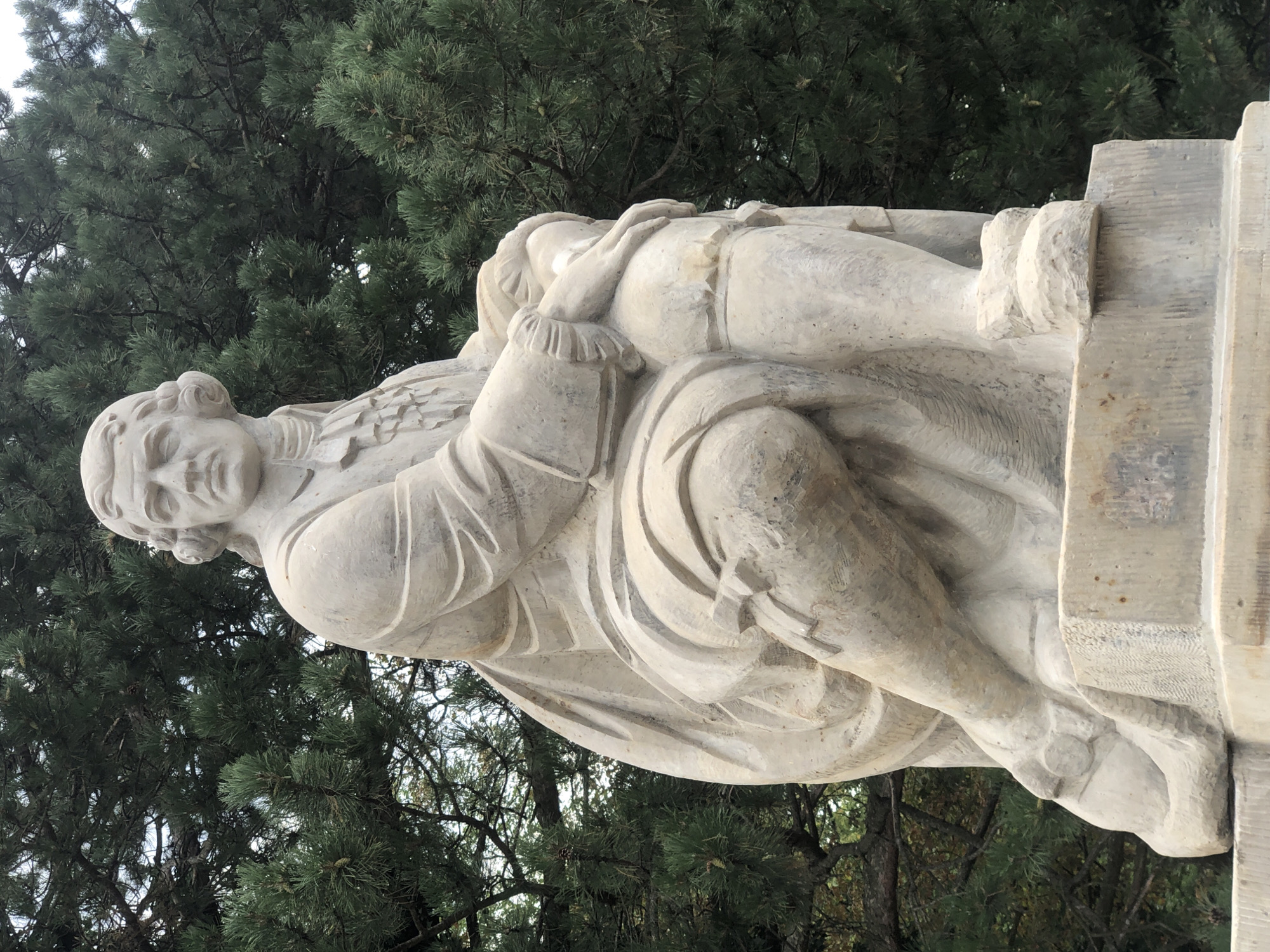 A full-body, eye-level, outdoor shot captures a light-colored stone statue of a man seated on a stone pedestal. The man has medium-length hair styled in curls, a long, collared coat, and breeches. His left leg is crossed over his right, and his left hand rests on his right knee. The statue is positioned in front of a backdrop of dark green pine trees. The statue appears to be located in Warsaw, Poland, specifically on Plac Bankowy, near the Zachęta National Gallery of Art, and is a monument to Stanisław Moniuszko.