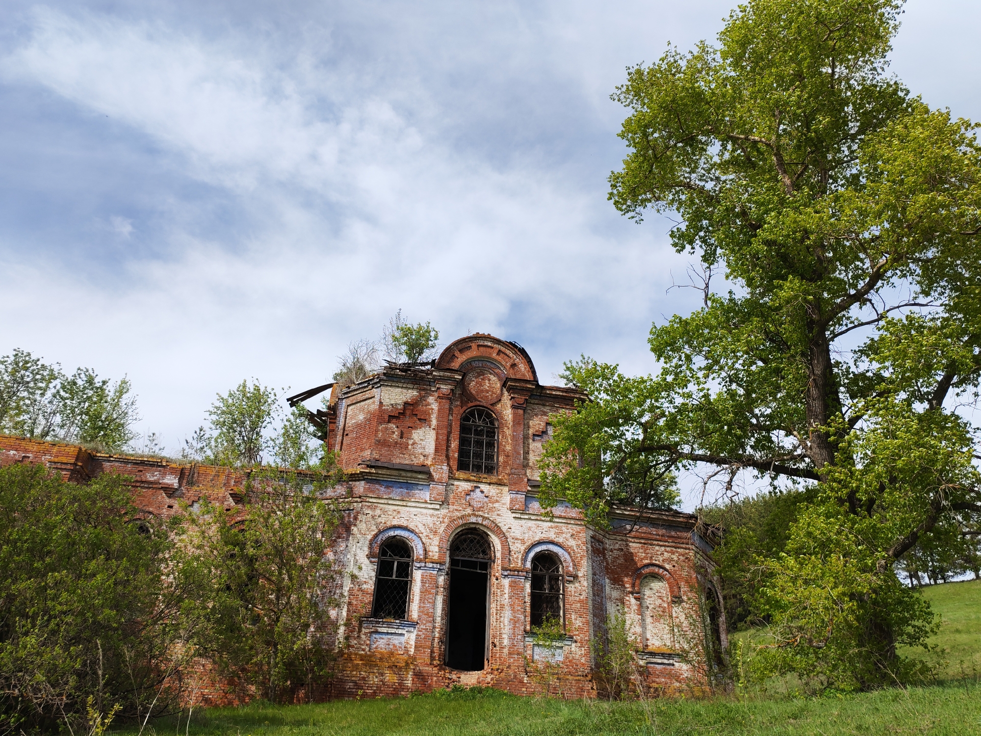 The image shows the ruined Church of the Life-Giving Trinity in the village of Крынды, Russia. The building is constructed of red brick and is heavily weathered, with missing sections of the roof and walls. Vegetation is growing on and around the structure. A large tree stands to the right of the building, partially obscuring the view. The sky is overcast with some patches of blue visible. The ground is covered in green grass.