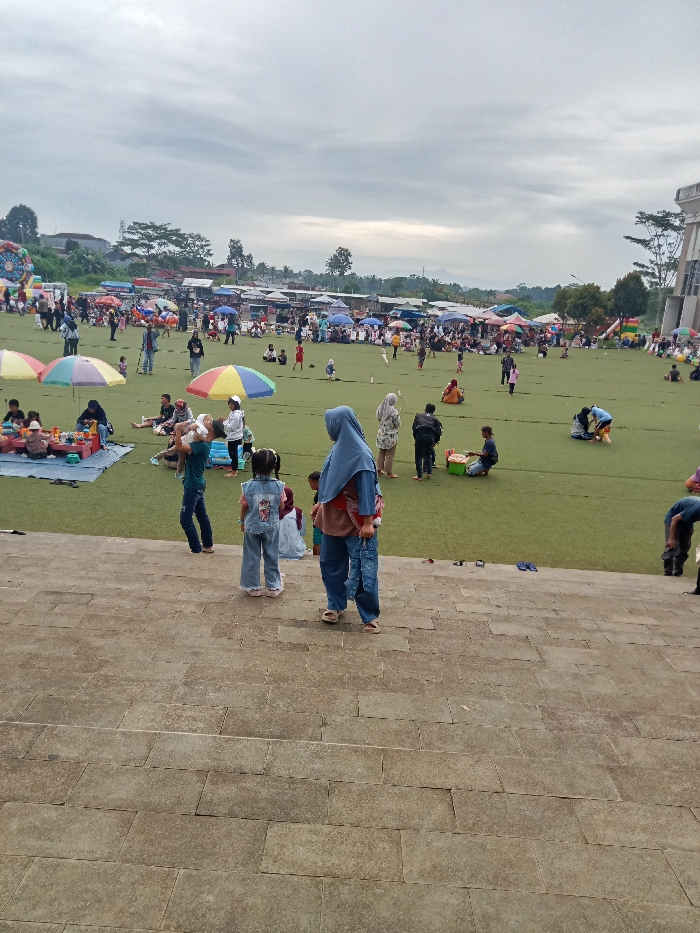 This image depicts a bustling outdoor gathering on a large, grassy field (likely artificial turf) in Singaparna, Indonesia, under an overcast sky. In the immediate foreground, wide, gray stone steps lead down into the scene.
The field is populated by numerous people of various ages, including many families with children and adults. People are seen relaxing, socializing, and picnicking; many are seated on mats or directly on the grass. Several colorful, market-style umbrellas are scattered across the field, providing shade. Children are visible playing, and an inflatable bounce house can be seen on the far left, indicating a family-friendly event.
Along the far edge of the field, numerous small market stalls and tents are set up in rows, suggesting a community fair, market, or festival. People are observed browsing these stalls. A vendor is visible near a small cart. On the far right, a modern-looking building with angled architectural elements stands.
The time of day appears to be late morning or afternoon, given the cloudy conditions which create a soft, even light. The overall mood is relaxed and communal, typical of a public gathering or recreational outing. Clothing choices, including hijabs worn by several individuals, align with the cultural context of Indonesia. No discernible text is visible in the image.