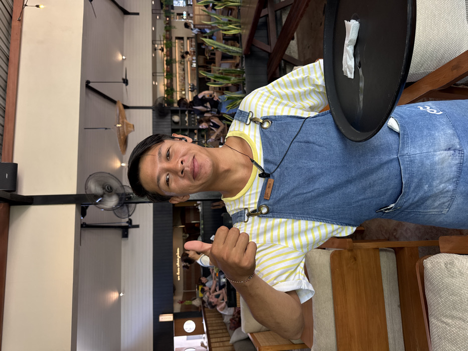 An indoor-outdoor dining establishment in Pecatu, Indonesia, is depicted. A male individual, appearing to be a server, occupies the foreground, facing the camera with a smile. He wears a yellow and white striped t-shirt under a denim bib apron secured with brass-colored buckles; a small brown patch is affixed to the apron's bib, and a wave-like logo is present on the lower right. A black wired earbud is inserted into his left ear, with the wire extending downwards. A silver-toned bracelet adorns his right wrist, and he holds a black serving tray in his left hand, which contains a crumpled white paper item. His right hand is raised, forming a 'thumbs up' gesture. A pen is visible in a pocket on his apron. The background reveals a spacious restaurant with high, angled ceilings featuring exposed dark beams, white paneling, multiple ceiling fans, and woven light fixtures. Seating arrangements include upholstered wooden chairs and booths with light cushions on the left, and wooden picnic-style tables and benches on the right, separated by planters containing snake plants. A bar area with bottles and hanging plants is visible in the mid-ground. Multiple patrons are seated throughout the establishment. The environment is brightly lit, suggesting daylight.