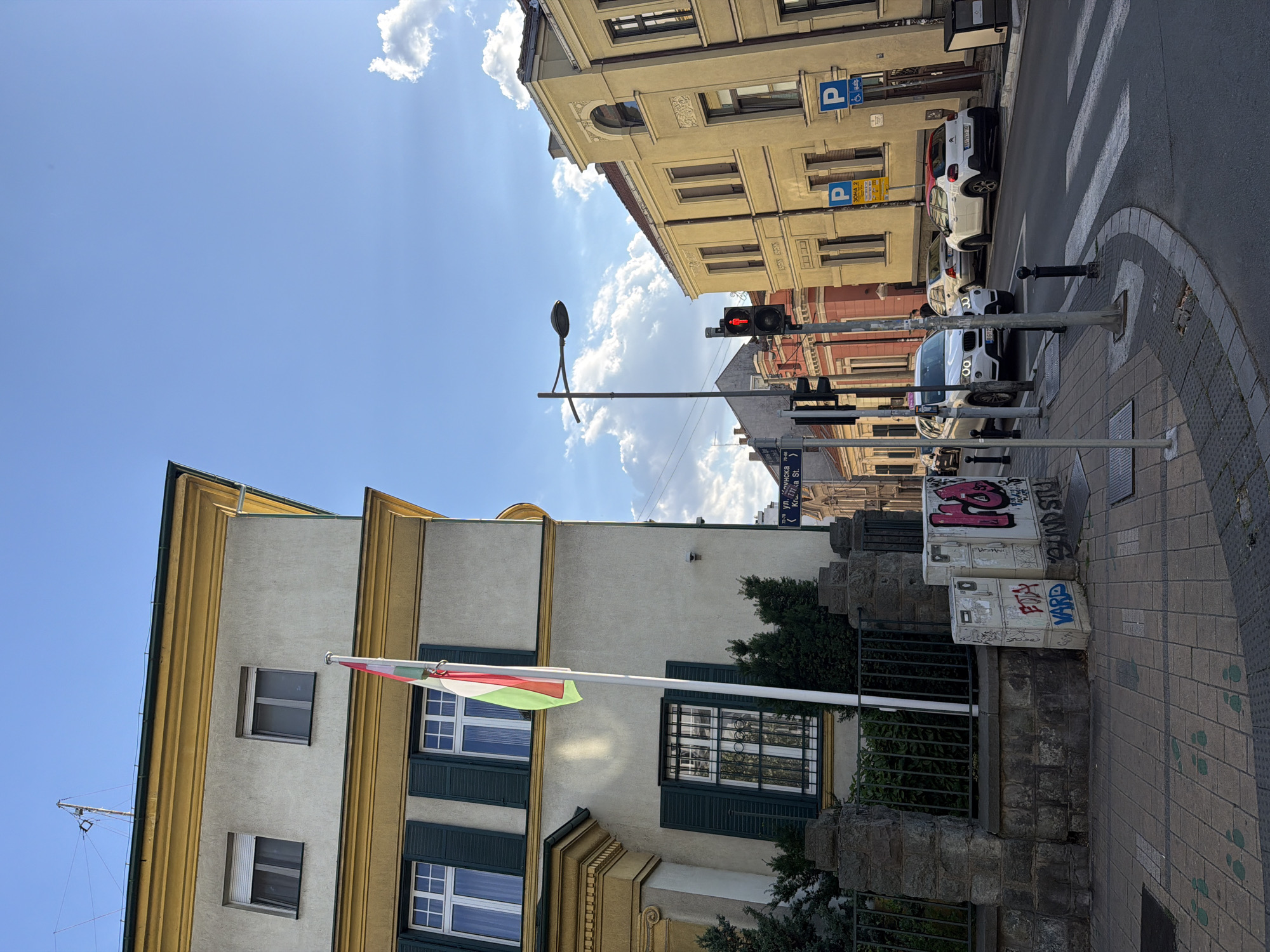 This media file depicts an outdoor scene in Vracar, Serbia, on a sunny day. The perspective is angled, looking up at buildings and the sky.
Prominently featured is a building with a light-colored facade and yellow decorative trim. Two stories are visible, each with multiple windows. The lower story has dark green shutters, and one window is barred. A flag with red, white, and green stripes is flying from a flagpole attached to the building.
Across the street, another building with a light beige facade displays yellow signs with blue parking symbols, indicating designated parking areas. A red traffic light is visible, suggesting an intersection. Several cars are parked along the street, including a white Citroën and a white BMW.
The street itself is paved with gray tiles and features a crosswalk marked with white stripes. A sidewalk with brick paving runs alongside the street. There are several lampposts and traffic signals. Graffiti is visible on a few white rectangular structures on the sidewalk.
The sky is bright blue with scattered white clouds, and sunlight streams through, casting shadows and highlights. The overall impression is a typical urban street scene during daylight hours. Some text is visible on street signs, including what appears to be Ukrainian street names and numbers, and graffiti on the white boxes.