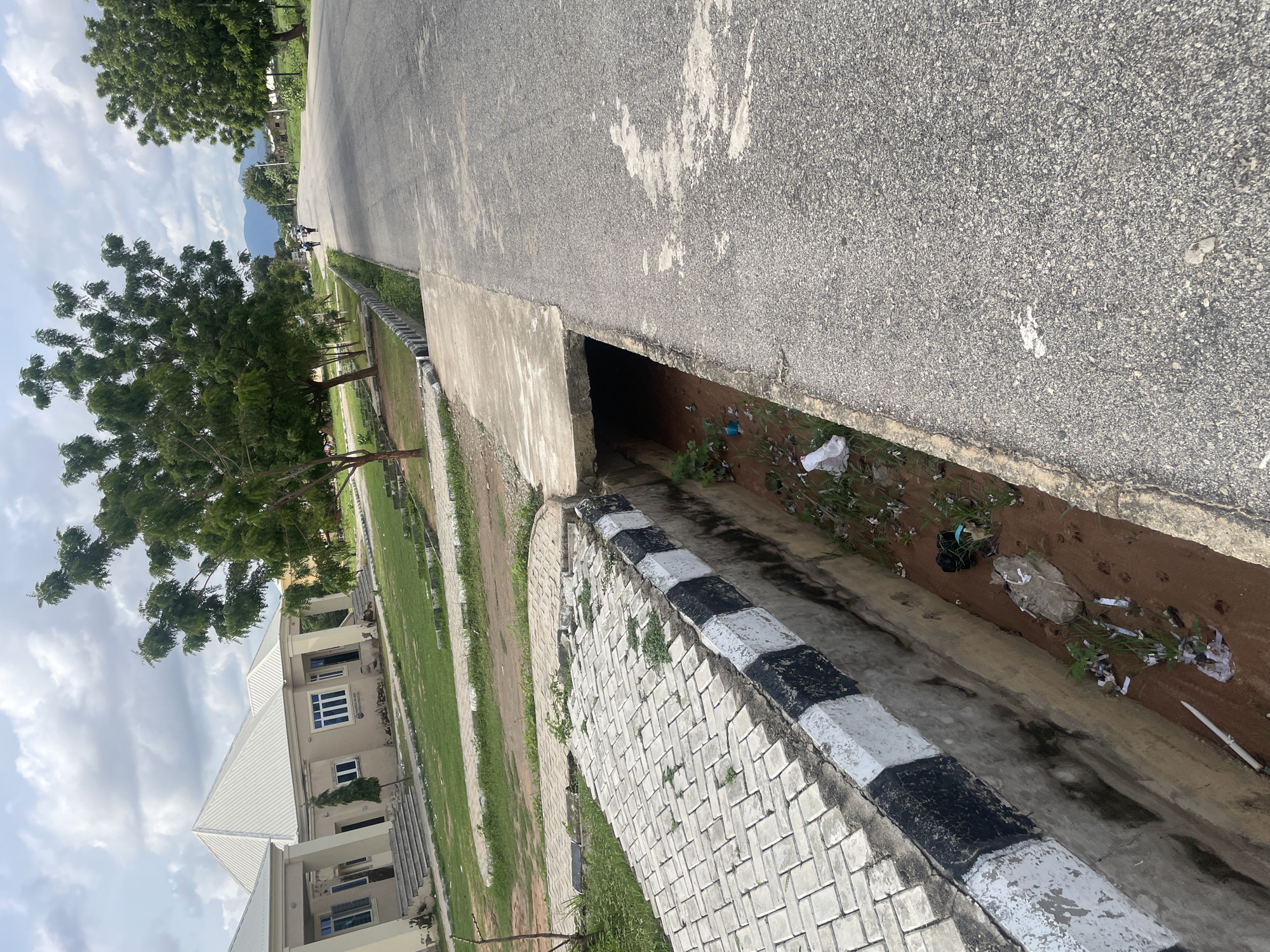 The image depicts a street scene in Jalingo, Nigeria, characterized by a concrete road running diagonally through the frame. On the left side of the road, a drainage ditch is visible, filled with reddish-brown soil, scattered debris including plastic bags and white paper, and some sparse vegetation. The edge of the ditch is lined with concrete, painted with alternating black and white stripes. Adjacent to the ditch is a paved walkway made of interlocking concrete blocks, also showing signs of wear and some overgrowth.
Further to the left, a light-colored building with a white, pitched roof is situated on a grassy lawn. This building appears to be a single-story structure with multiple windows and a covered entrance. Several trees with green foliage are scattered across the landscape, with one prominent tree in the mid-ground.
On the right side of the road, the asphalt surface shows some patching and wear. In the distance, the road continues, lined by trees and some structures. A few people are walking on the road in the distance, and two individuals are on bicycles. The sky is partly cloudy, with white clouds against a light blue background. The overall impression is that of an outdoor environment, possibly a residential or institutional area, with evidence of some littering.