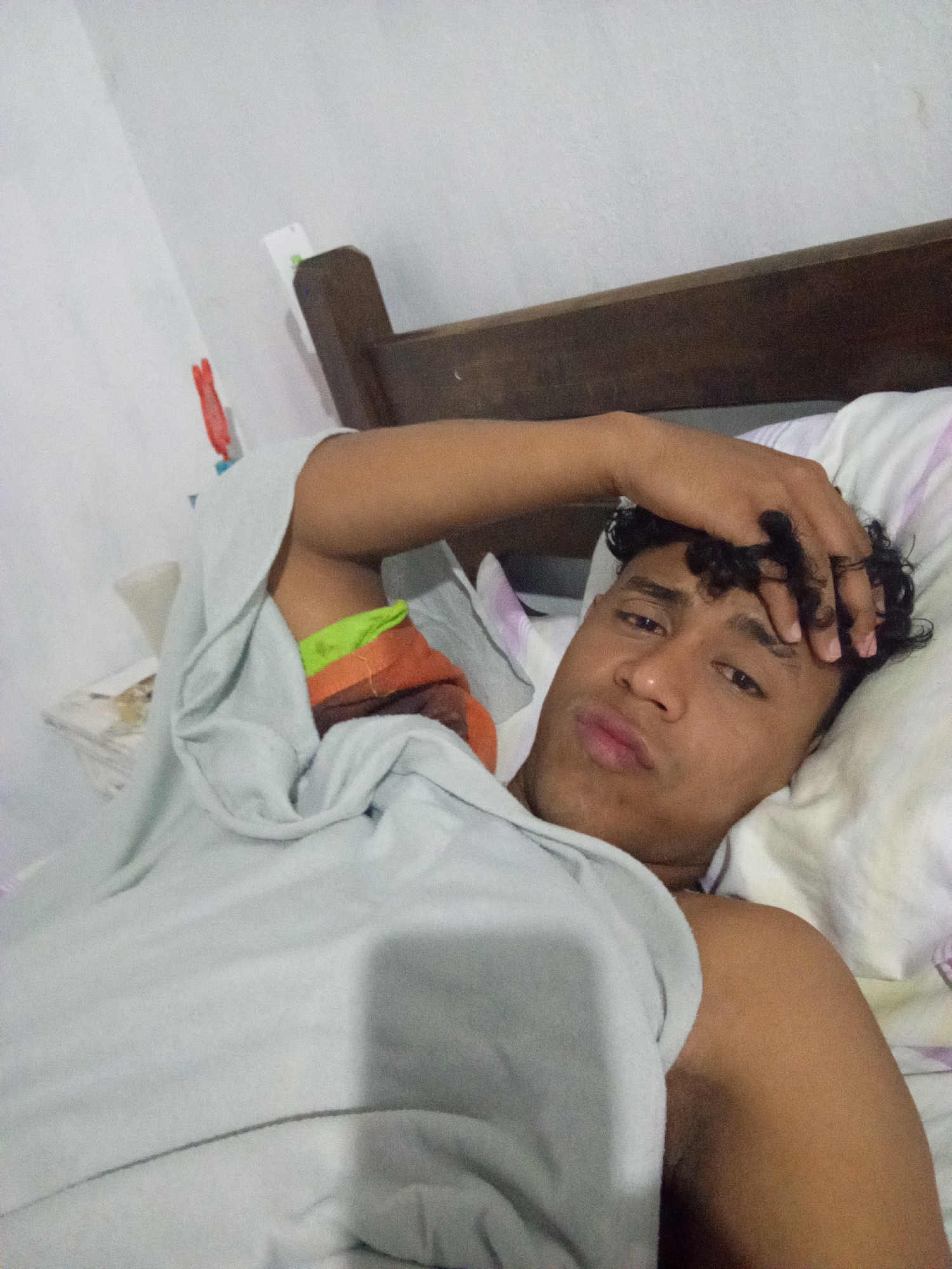 A young man with curly black hair is lying in a bed. He is partially covered by a light grey blanket, exposing his bare right shoulder. His left hand is resting on his forehead. The bed has a dark wood headboard and a white pillow with faint stripes. The background includes a white wall. The location is Macho Muerto, Venezuela.