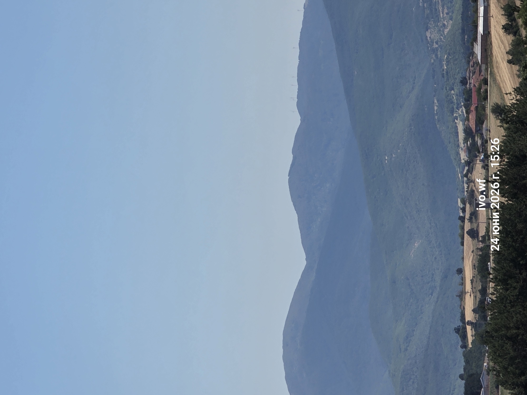 This media file displays a panoramic view of a mountainous landscape in Chal, Bulgaria. The scene is dominated by rolling, verdant hills that stretch across the frame, with a clear, pale blue sky above. In the distance, silhouetted against the horizon, are several wind turbines.
Towards the right side of the image, a village is nestled at the base of the mountains. Houses with red-tiled roofs are visible, interspersed with trees and some agricultural fields. A road runs alongside the village, and faint signs of human activity, possibly vehicles, can be observed. The overall setting suggests a rural and tranquil environment.
The image appears to be taken during the daytime, likely in the late afternoon or early evening, given the soft lighting. The weather seems clear and calm.
Visible text on the right side of the image reads: "ivo.wf 24 юни 2026 г., 15:26". This indicates the filename and the date and time the image was captured in Bulgarian.