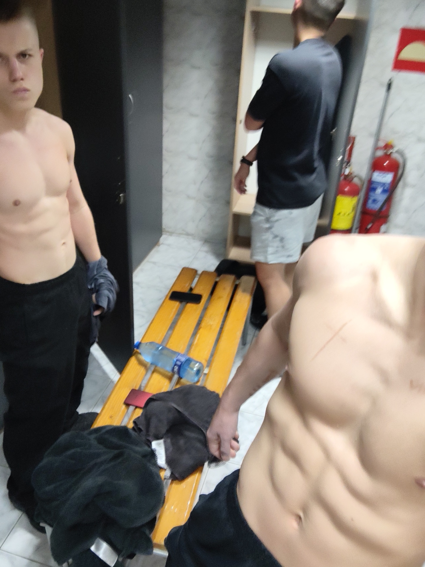 A brightly lit changing room contains three men. Two men are shirtless and wearing black bottoms, revealing toned torsos. One stands to the left, and the other is nearer to the camera on the right, with an abrasion across his upper chest. A third man is in the background with his back to the camera, wearing a black t-shirt and gray shorts. He stands next to open lockers.
A yellow wooden bench is in the center of the image. It contains a water bottle, dark towels, and what appears to be a mobile phone.
Two fire extinguishers are mounted to the wall next to the background man. The wall and floor are tiled. A partially visible red sign is hung near the lockers. The room appears to be in a gym in Kozloduy, Bulgaria.