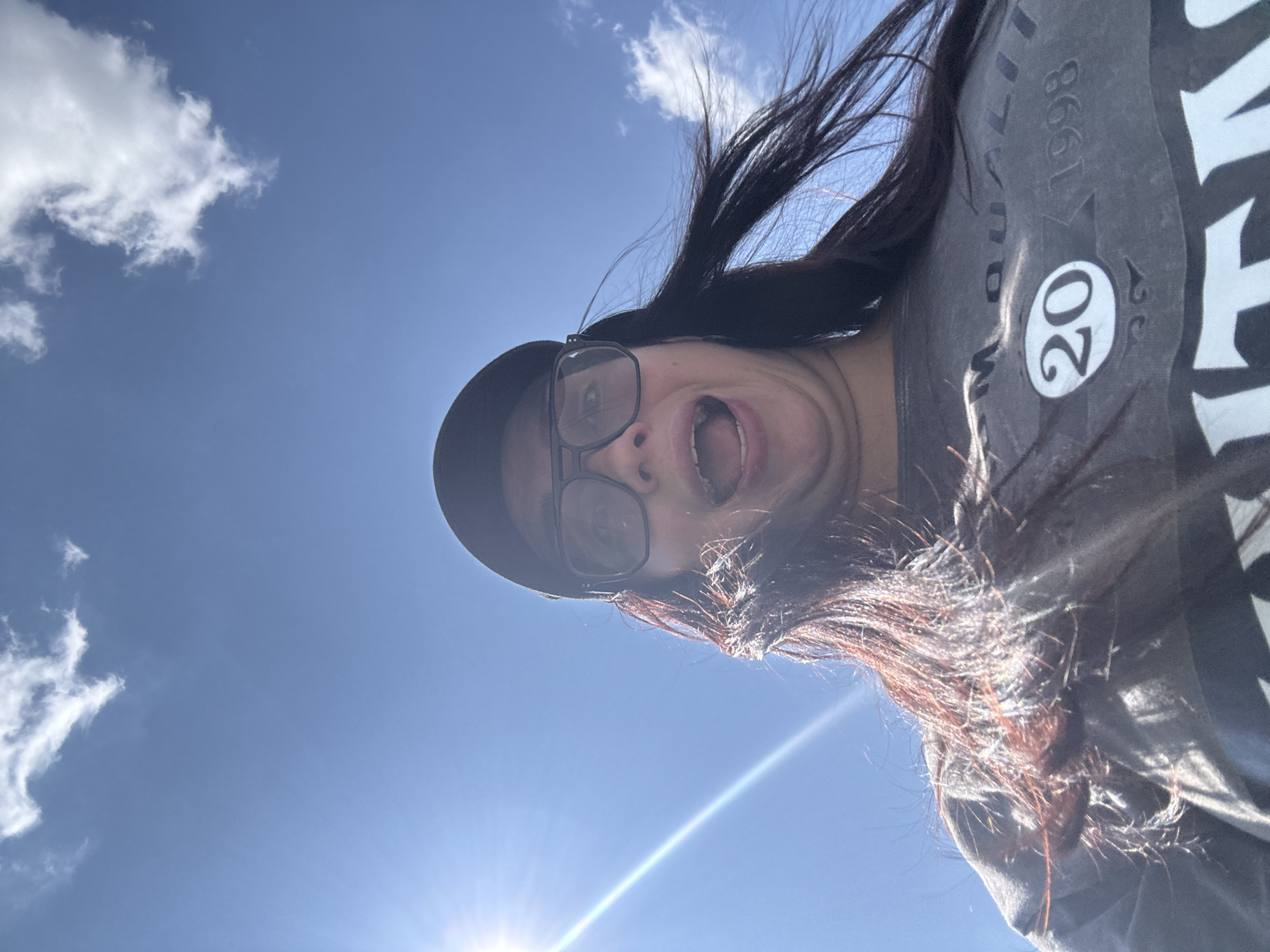 A low-angle outdoor shot captures a woman wearing a black baseball cap and large black-rimmed glasses. Her mouth is open in an exaggerated expression, and her dark hair is being blown by the wind. She is wearing a dark gray t-shirt with faded text that includes the numbers "20" and "1998". The background is a bright blue sky with a few wispy clouds and a sun flare in the upper left corner. There is no discernible information in the image to suggest a location in Tolyatti, Russia.
