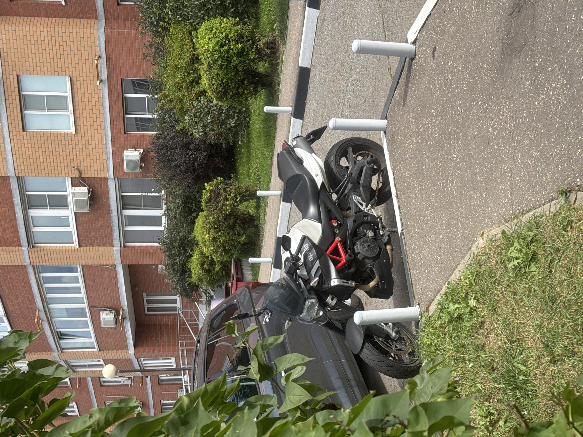 In the city of Sovkhoz imeni Lenina, Russia, a black and white Aprilia motorcycle is parked on a paved surface adjacent to a brick apartment building. The motorcycle is positioned on a marked parking area with white bollards. A portion of a dark-colored car is visible to the left of the motorcycle, partially obscured by lush green foliage. The background features the multi-story brick building with numerous windows and air conditioning units. Green bushes and grass surround the parking area and the building. The scene appears to be a typical urban residential environment on a sunny day.