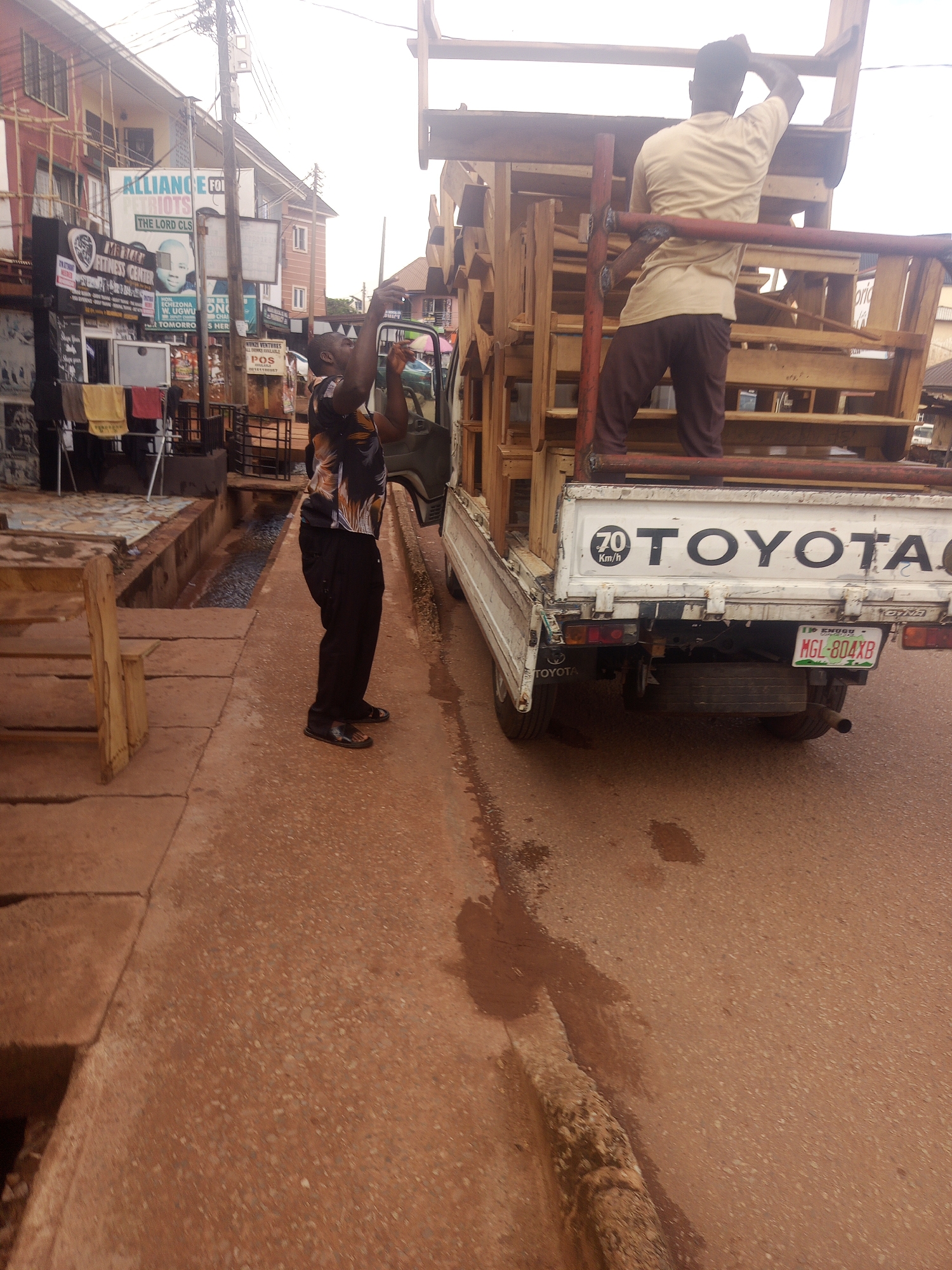 This image depicts a scene on an urban street in Enugu, Nigeria, likely near a commercial area judging by the buildings and signage. In the foreground, a drainage channel runs along the side of the road, bordered by concrete structures. A man in a patterned shirt and dark pants is standing on the road, facing away from the camera and looking upwards, with his arms raised as if adjusting something. He is positioned near the rear of a white Toyota truck. The truck is loaded with a large wooden structure, possibly crates or scaffolding, made of light-colored wood and red metal poles. The truck's tailgate is visible, with "TOYOTA" branding and a license plate "MGL-804XB" also visible. Behind the man and truck, buildings with multiple stories line the street, featuring various signs including "ALLIANCE FOR THIS THE LORD CHOOSES," "FITNESS CENTER," and others in local languages and English. Laundry is hanging on a rack in front of one of the buildings. The road surface is paved and appears somewhat wet or dusty.